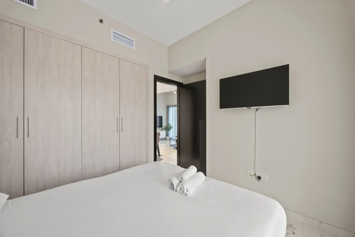 Modern bedroom with tall light-wood wardrobes along left wall and a wall-mounted TV on the right; bed in foreground with rolled towels.