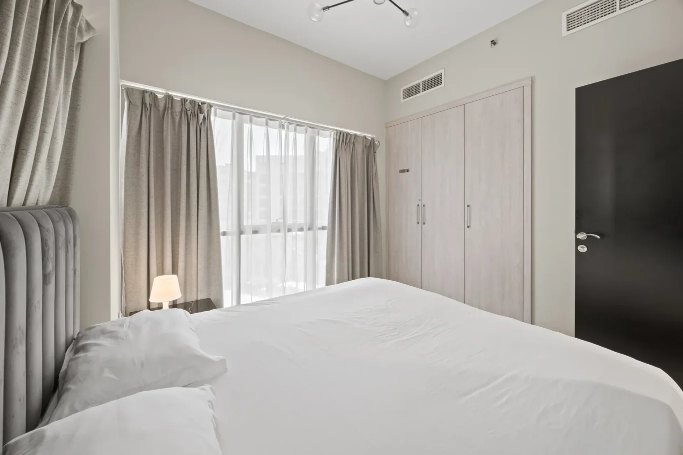 Hotel bedroom with white bedding, grey headboard, and large window with sheer and blackout curtains.