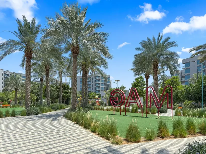 Red metal sculpture spelling 'MAC' on a grassy plaza bordered by palm trees in a sunny residential courtyard.