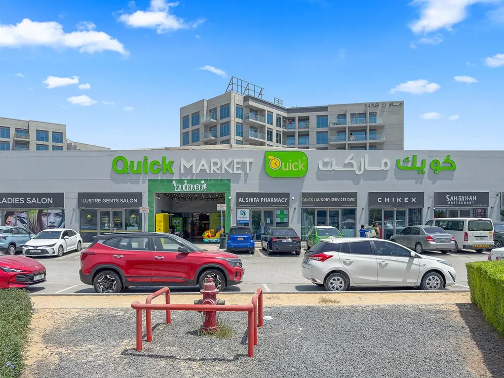 Exterior view of a Quick Market storefront with green logo and Arabic text, cars parked in front under a blue sky.