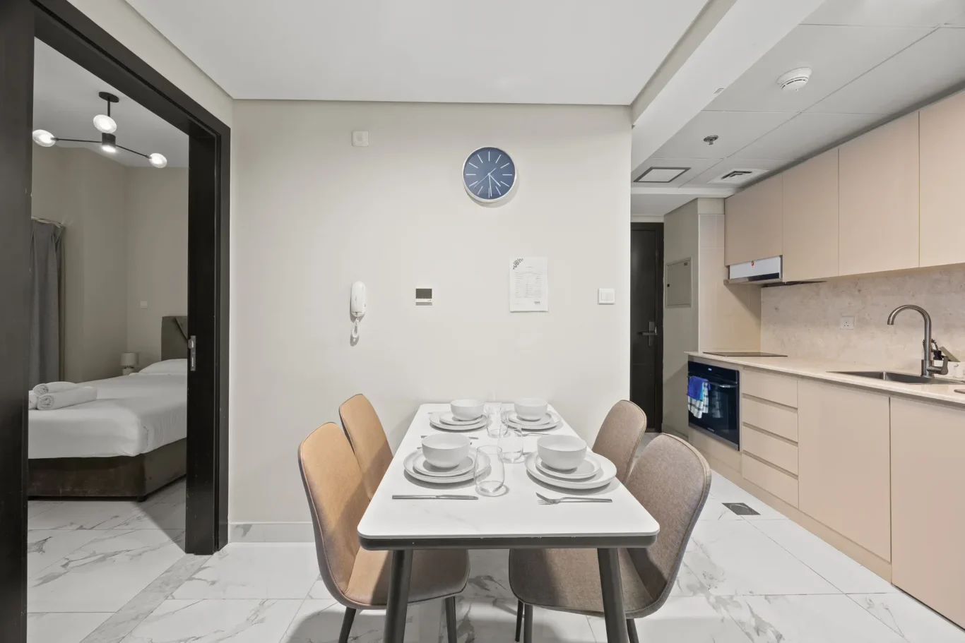 Dining table set for six in a modern kitchen, beige chairs, and a wall clock on the central wall; bedroom door visible on the left.
