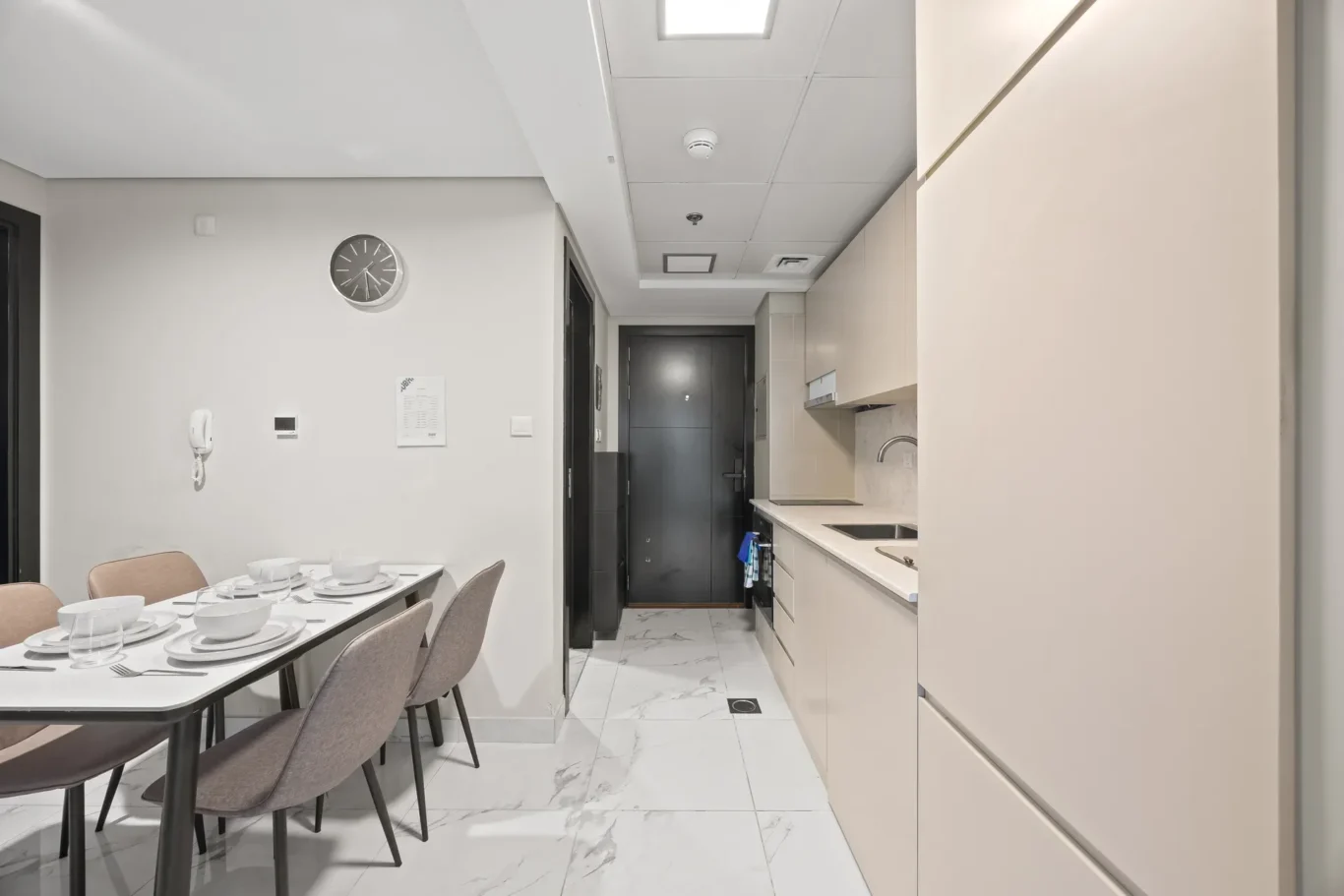 Modern galley kitchen and dining area with a table set for six, beige chairs, wall clock, and white cabinetry along the right edge.