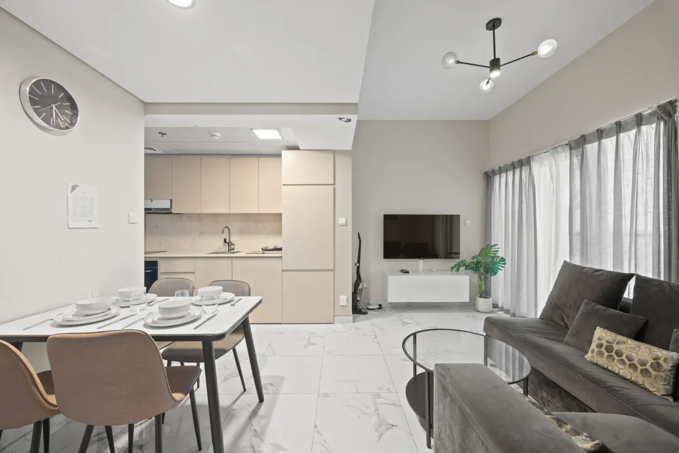 Open-plan living area with a beige kitchen, a dining table set, and a gray sofa facing a wall-mounted TV.