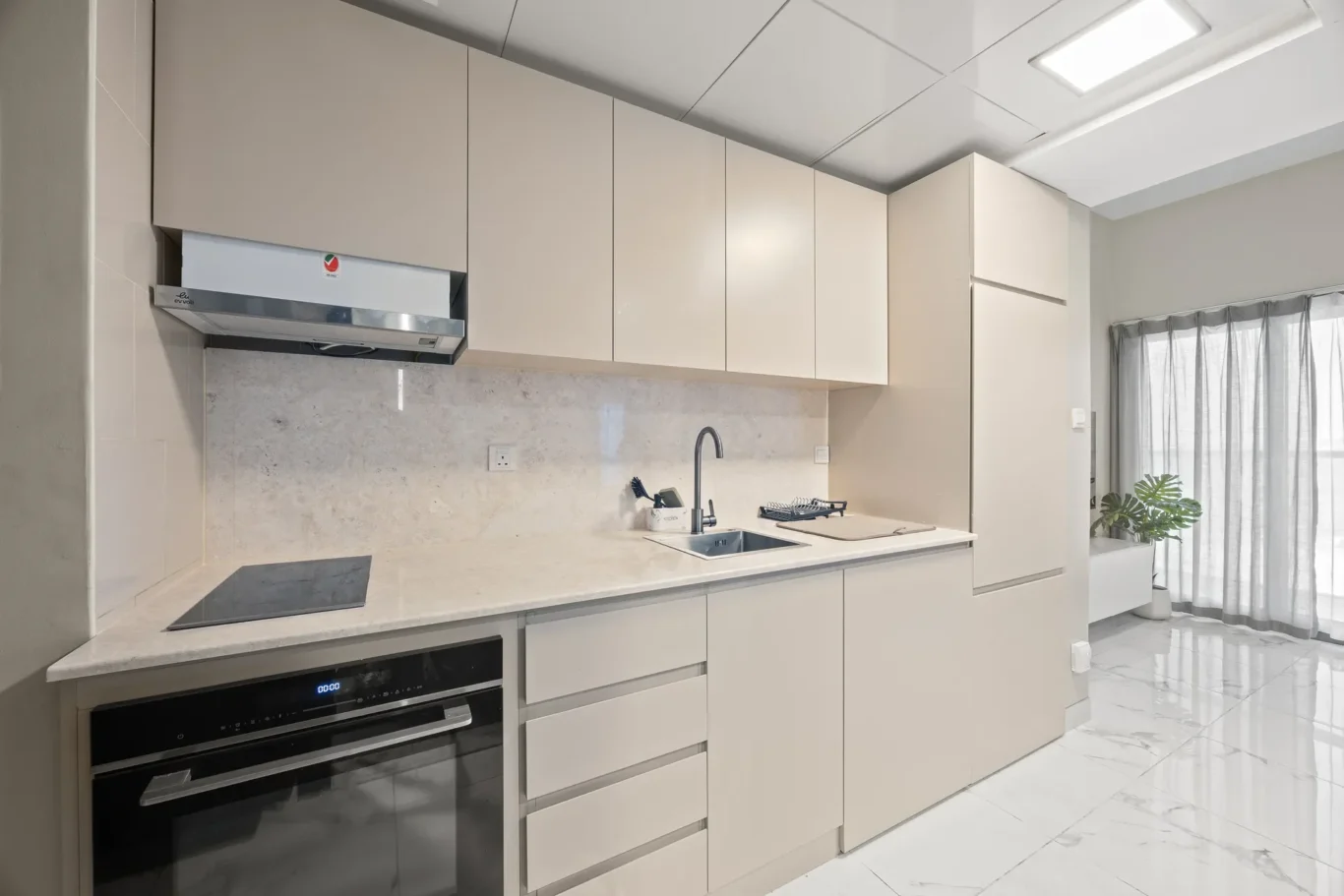 Modern beige kitchen with built-in oven, induction cooktop, range hood, and marble countertops.