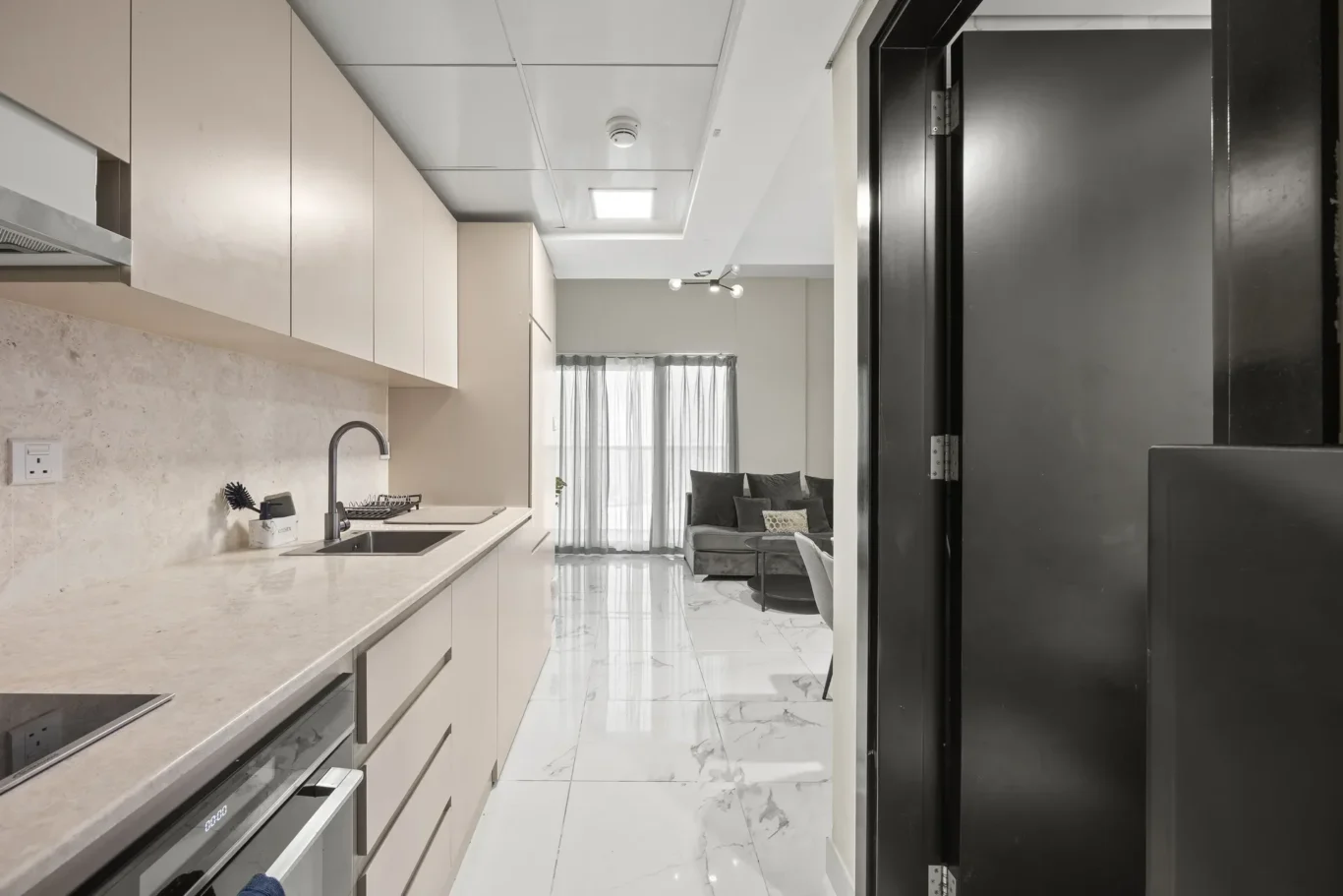 Modern galley kitchen with white cabinets, marble countertop, and sink; open living area with sofa visible beyond.