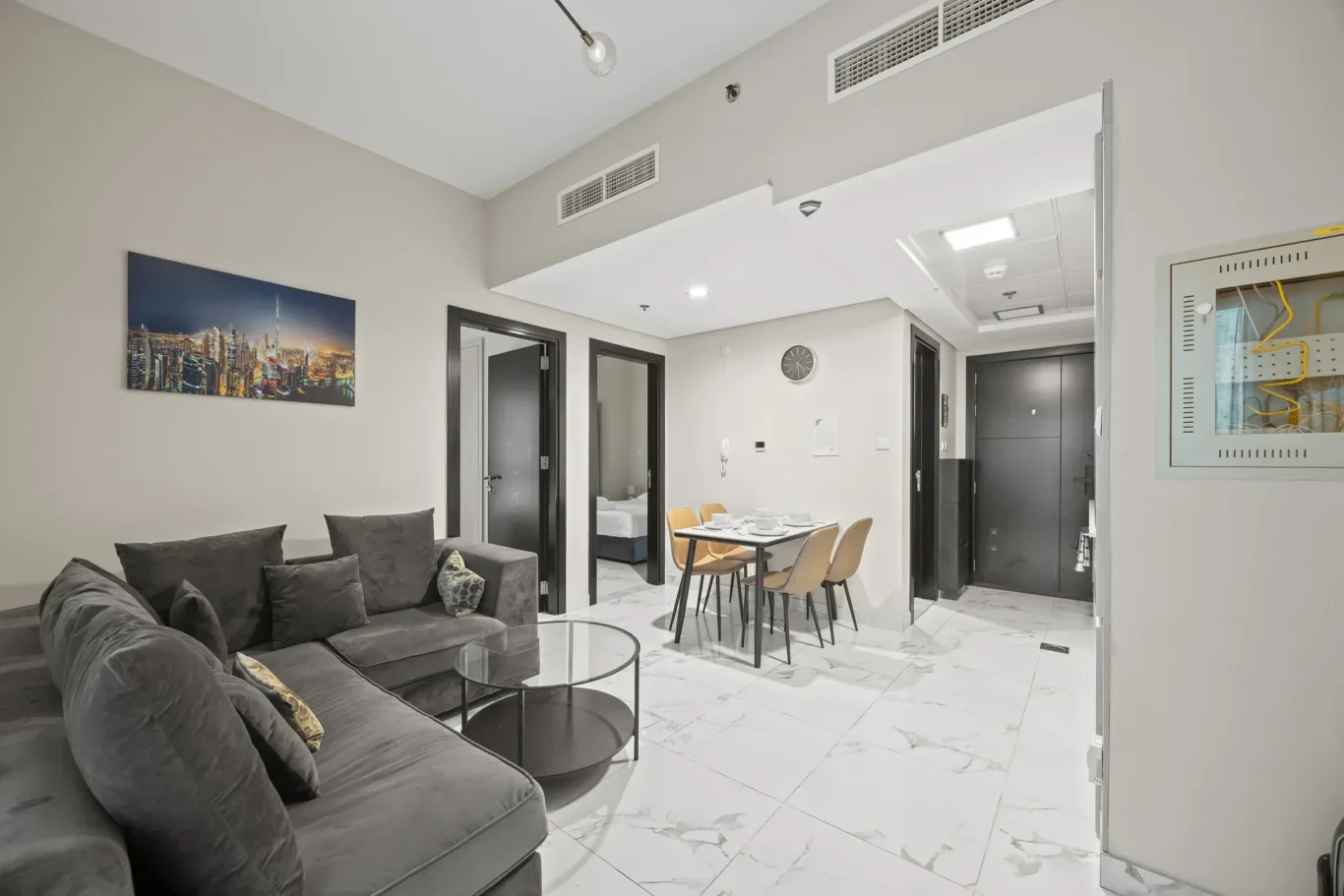 Living area in a modern apartment: gray sectional sofa with cushions, two round glass coffee tables, and a dining table with four chairs on a marble floor, plus a cityscape wall art.
