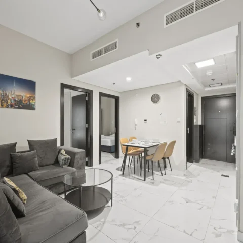 Living area in a modern apartment: gray sectional sofa with cushions, two round glass coffee tables, and a dining table with four chairs on a marble floor, plus a cityscape wall art.