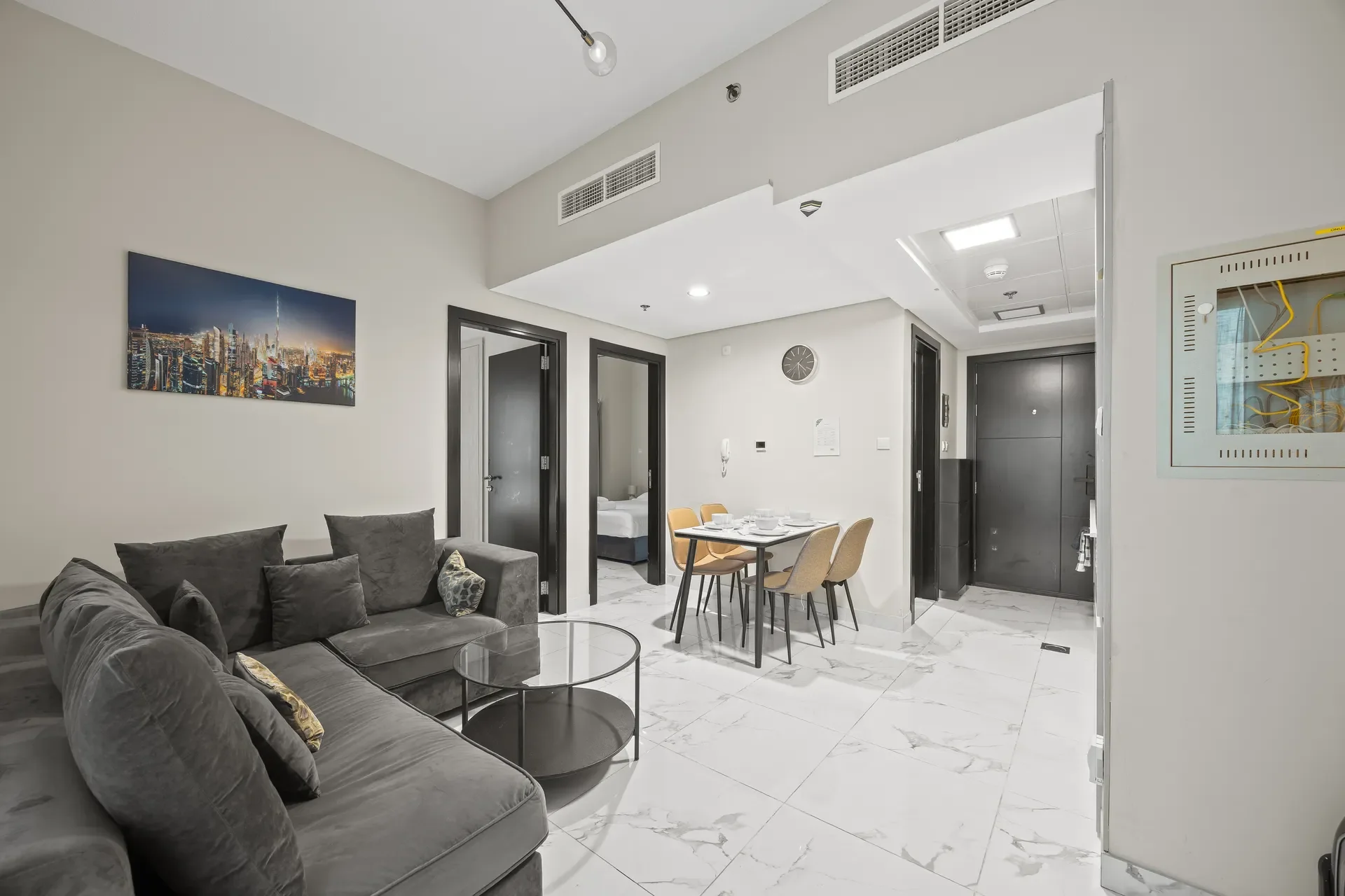 Living area in a modern apartment: gray sectional sofa with cushions, two round glass coffee tables, and a dining table with four chairs on a marble floor, plus a cityscape wall art.
