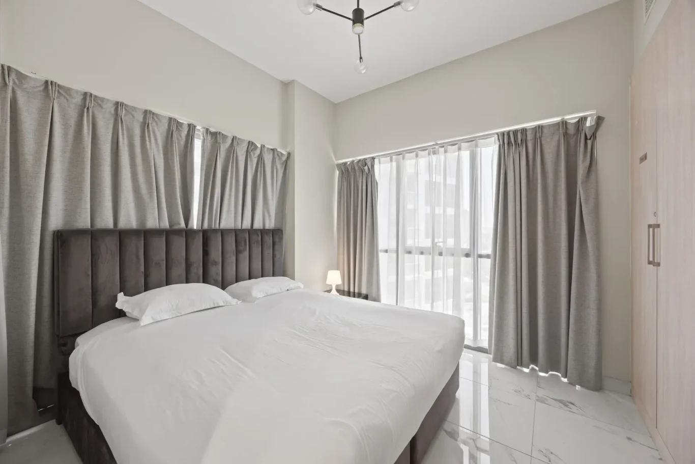 Bright modern bedroom with a large gray upholstered headboard, white linens, and marble-like floor; windows have sheer white and gray curtains.