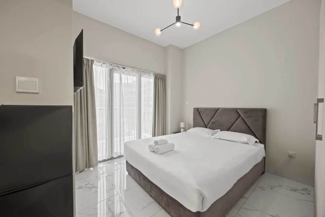 Hotel bedroom with a double bed, white linens, rolled towels, gray upholstered headboard, and marble floor.