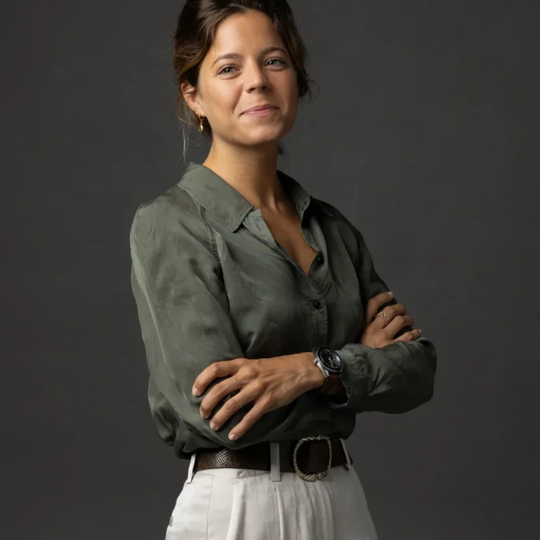 Portrait of a woman with dark hair in a green button-up shirt and white pants, arms crossed, against a dark gray backdrop.