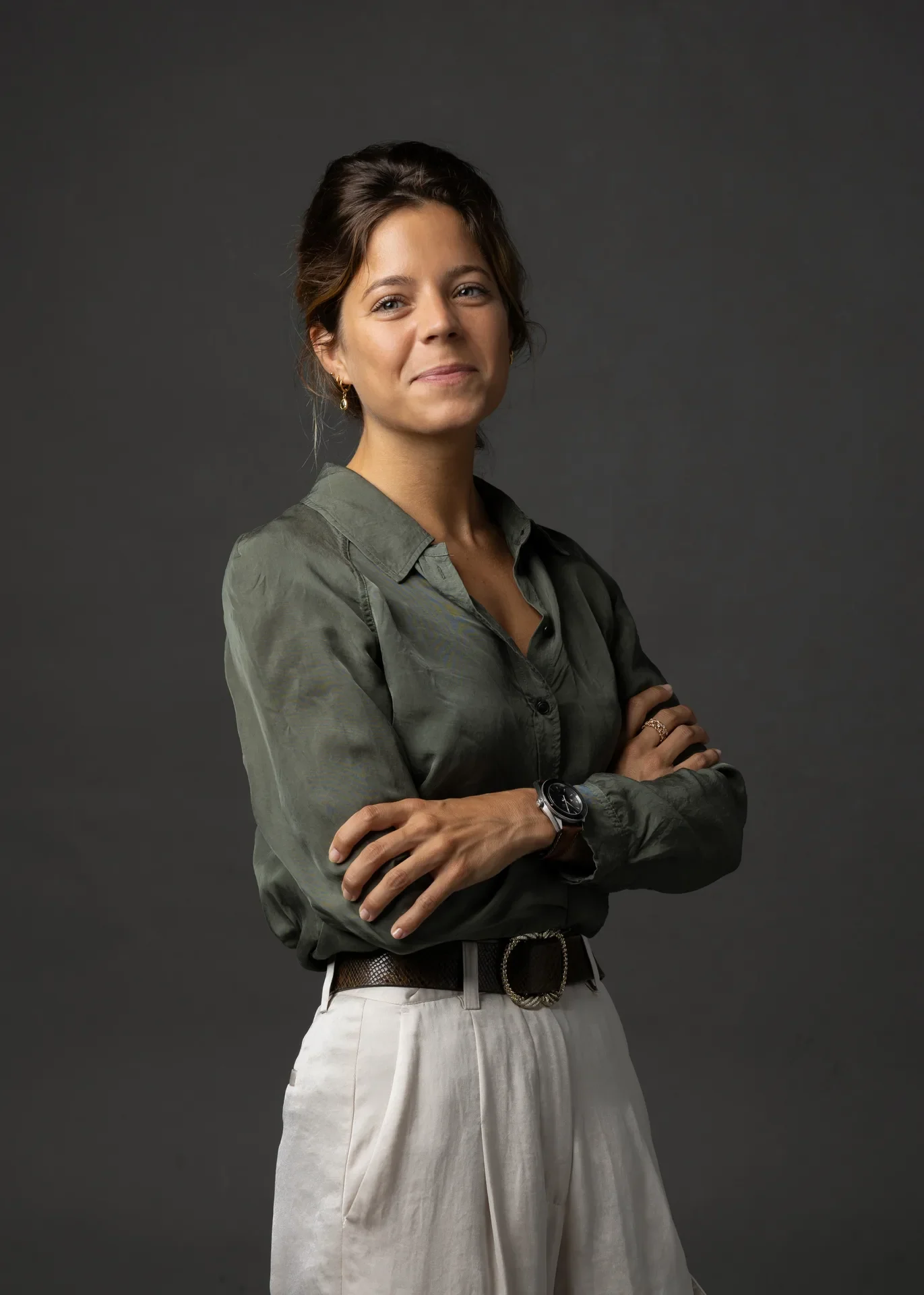 Portrait of a woman with dark hair in a green button-up shirt and white pants, arms crossed, against a dark gray backdrop.