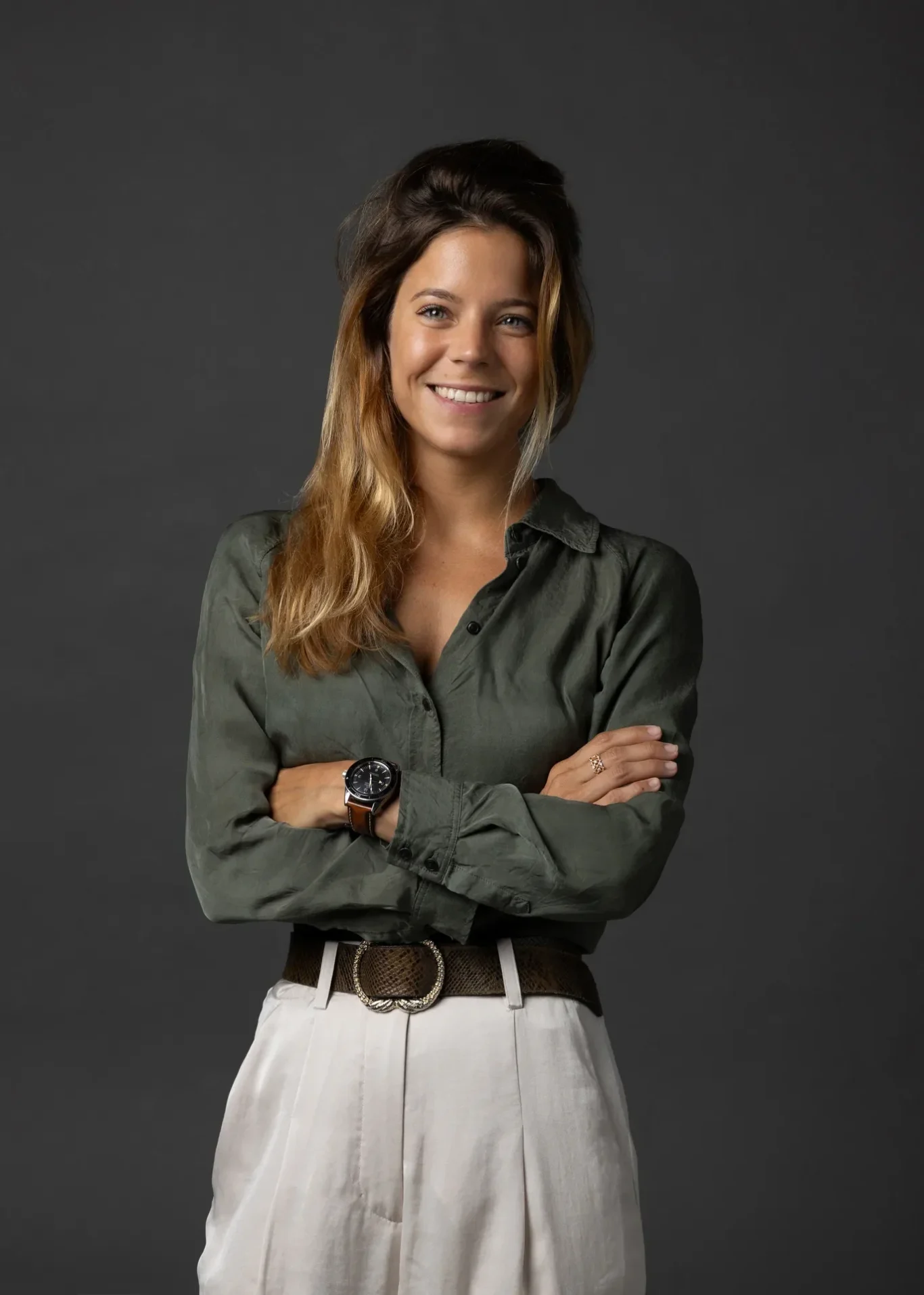 Smiling woman with arms crossed, wearing an olive green button-down shirt, beige belt, and light pants against a dark gray background.