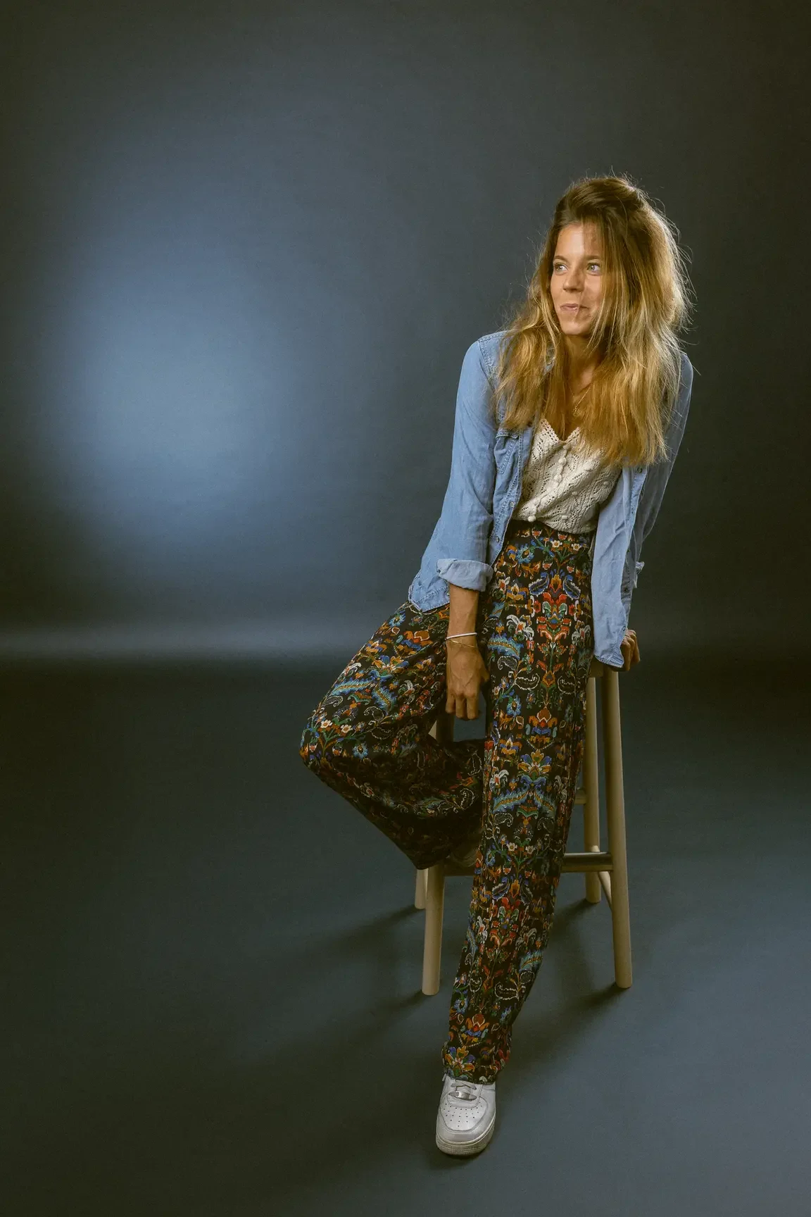 Young woman sits on a wooden stool in a studio, wearing a light denim shirt over a white lace top and colorful patterned pants, with white sneakers.