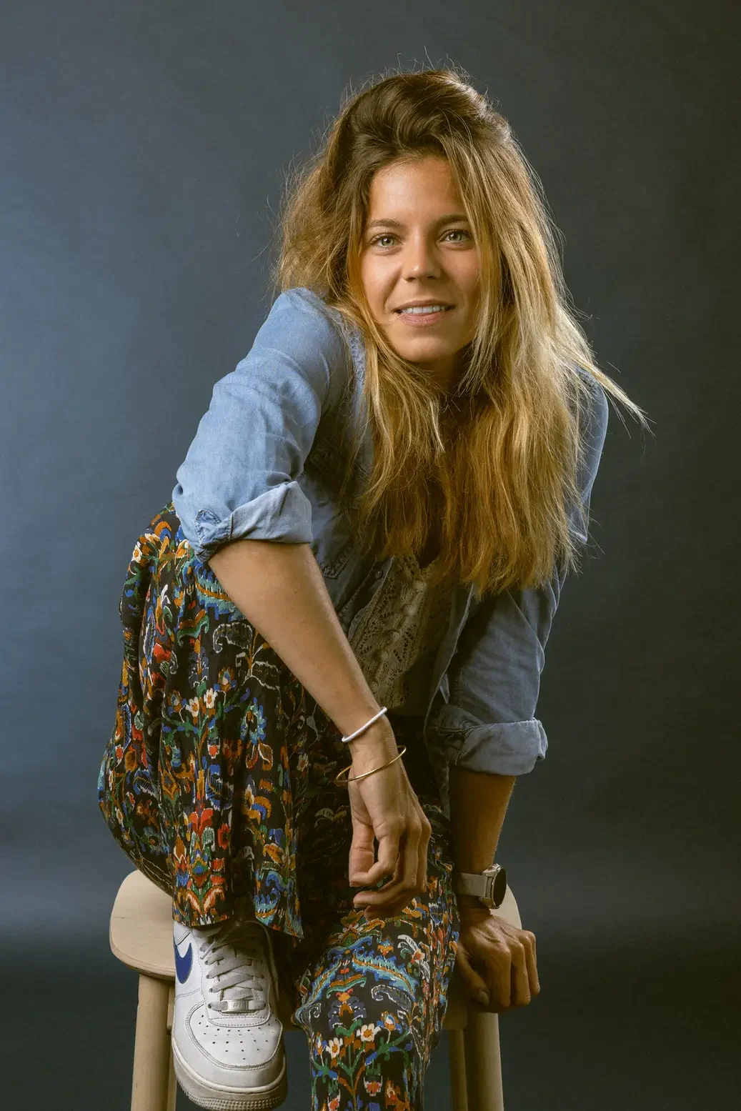 Young woman seated on a light stool, leaning forward with a warm smile, wearing a denim shirt and colorful patterned pants with white sneakers.