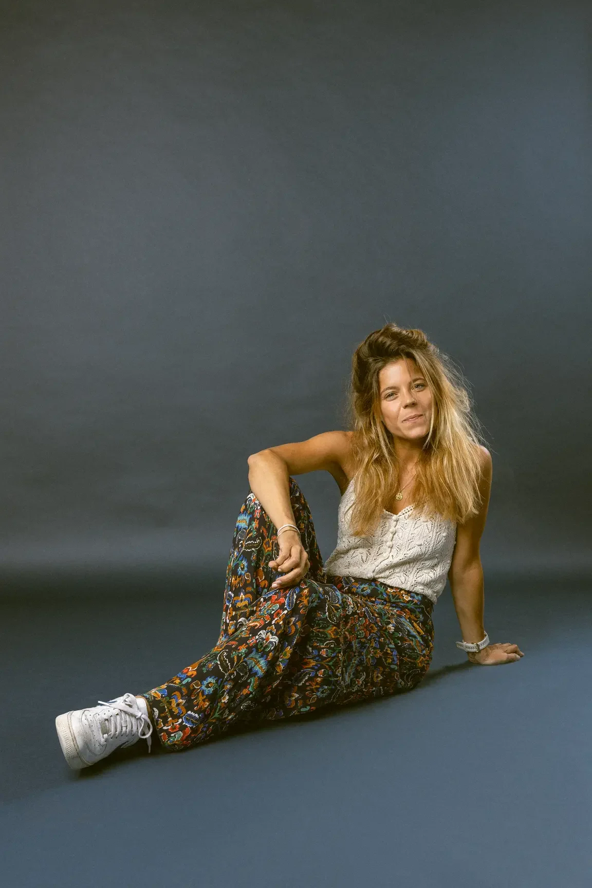 Woman seated on a dark backdrop, leaning on one hand with legs extended, wearing a white crochet top and colorful patterned pants, white sneakers.