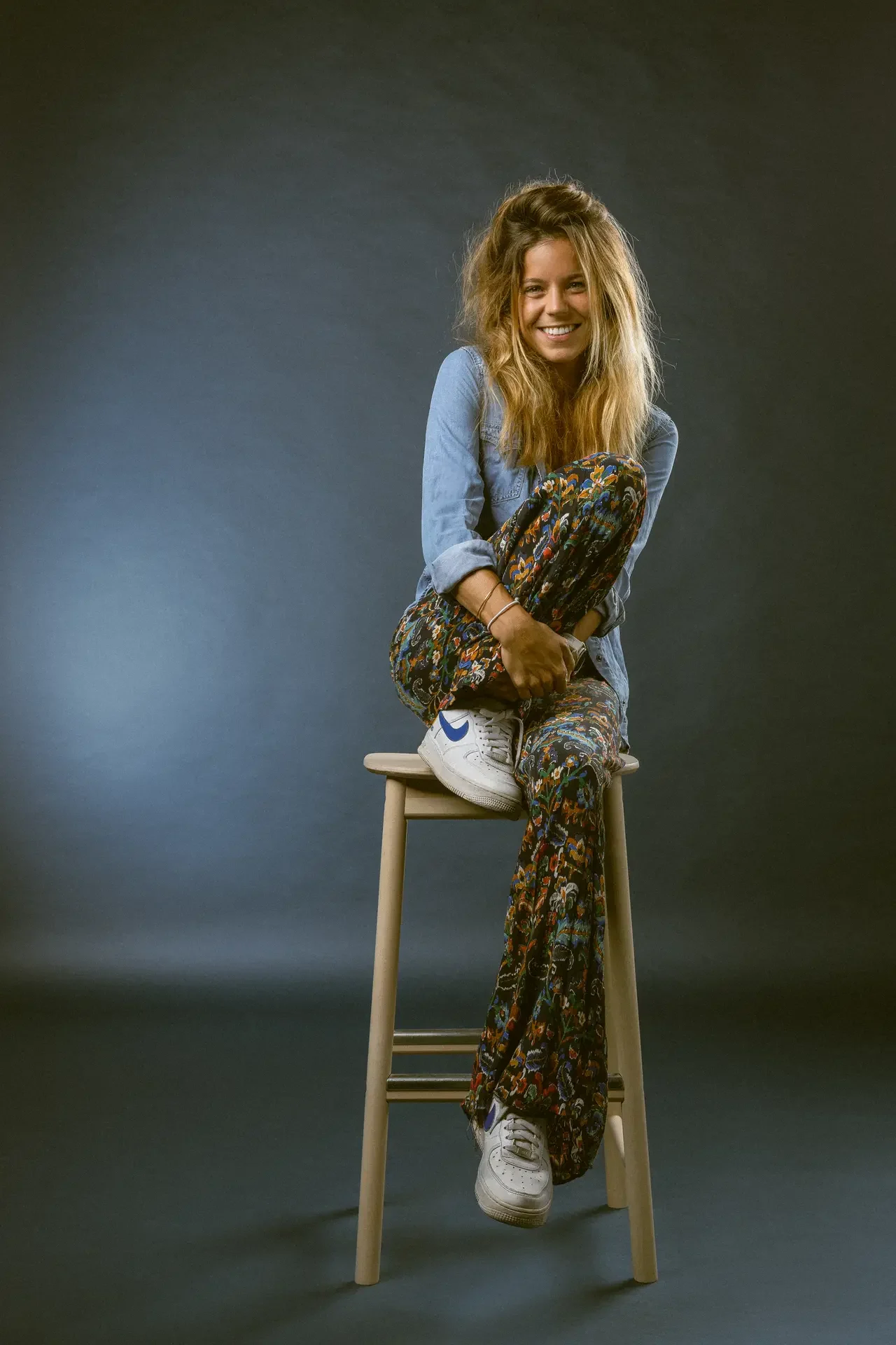 Smiling woman sits on a wooden stool with legs crossed, wearing a denim shirt, colorful patterned pants, and white sneakers against a dark studio backdrop.