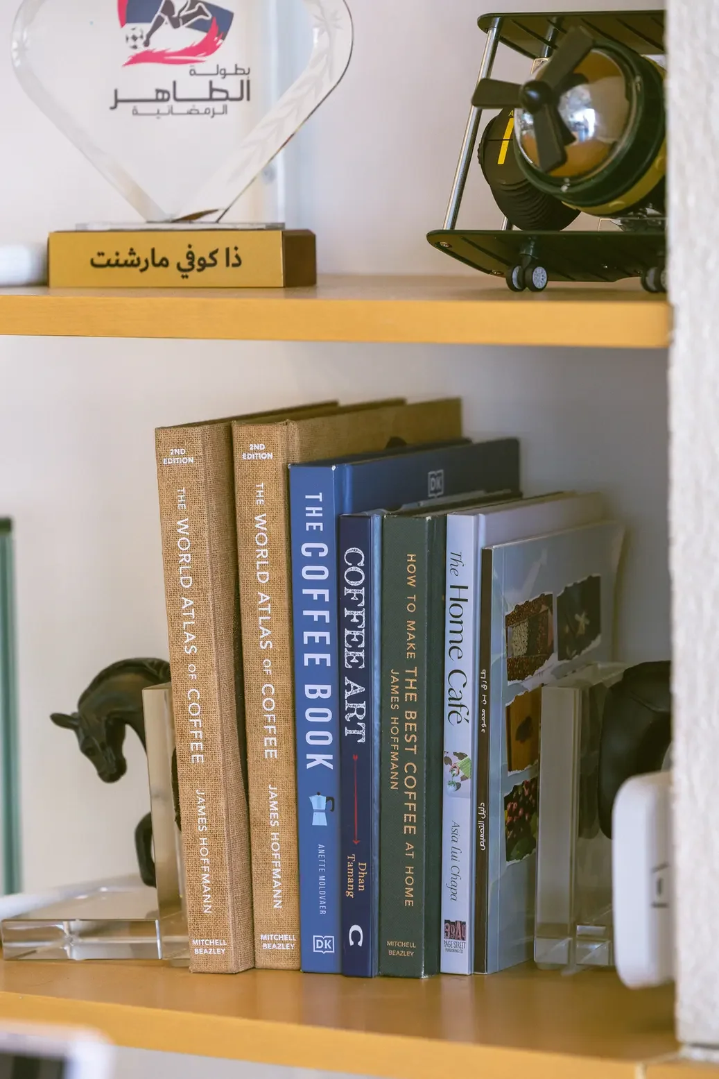 Shelf of coffee books, including The World Atlas of Coffee and The Coffee Book, with a glass trophy and a vintage diving helmet on top shelf.