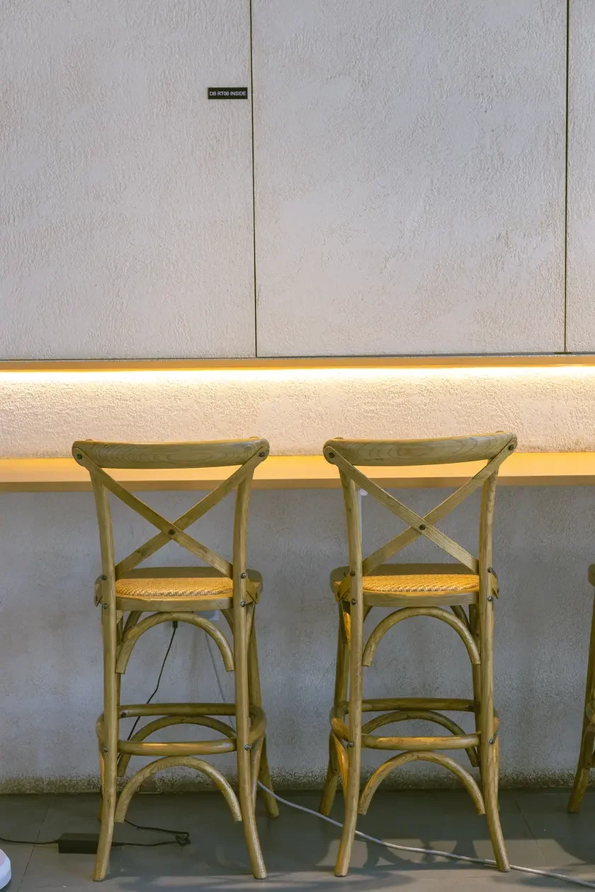 Two light-woven rattan bar stools sit at a glowing counter against a white textured wall in a cafe setting.