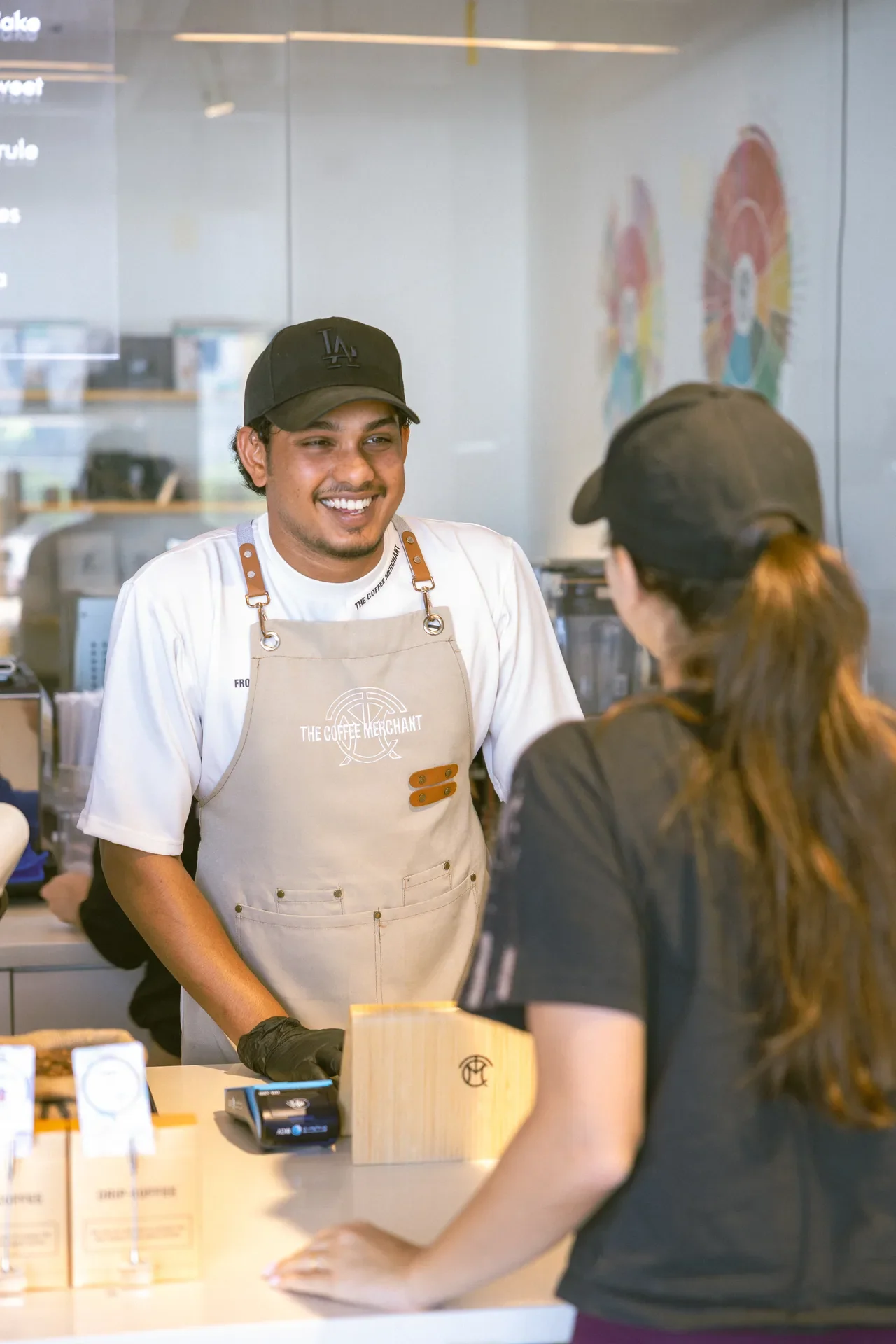 Smiling barista wearing a beige apron and black LA cap serves a customer at a coffee shop counter.