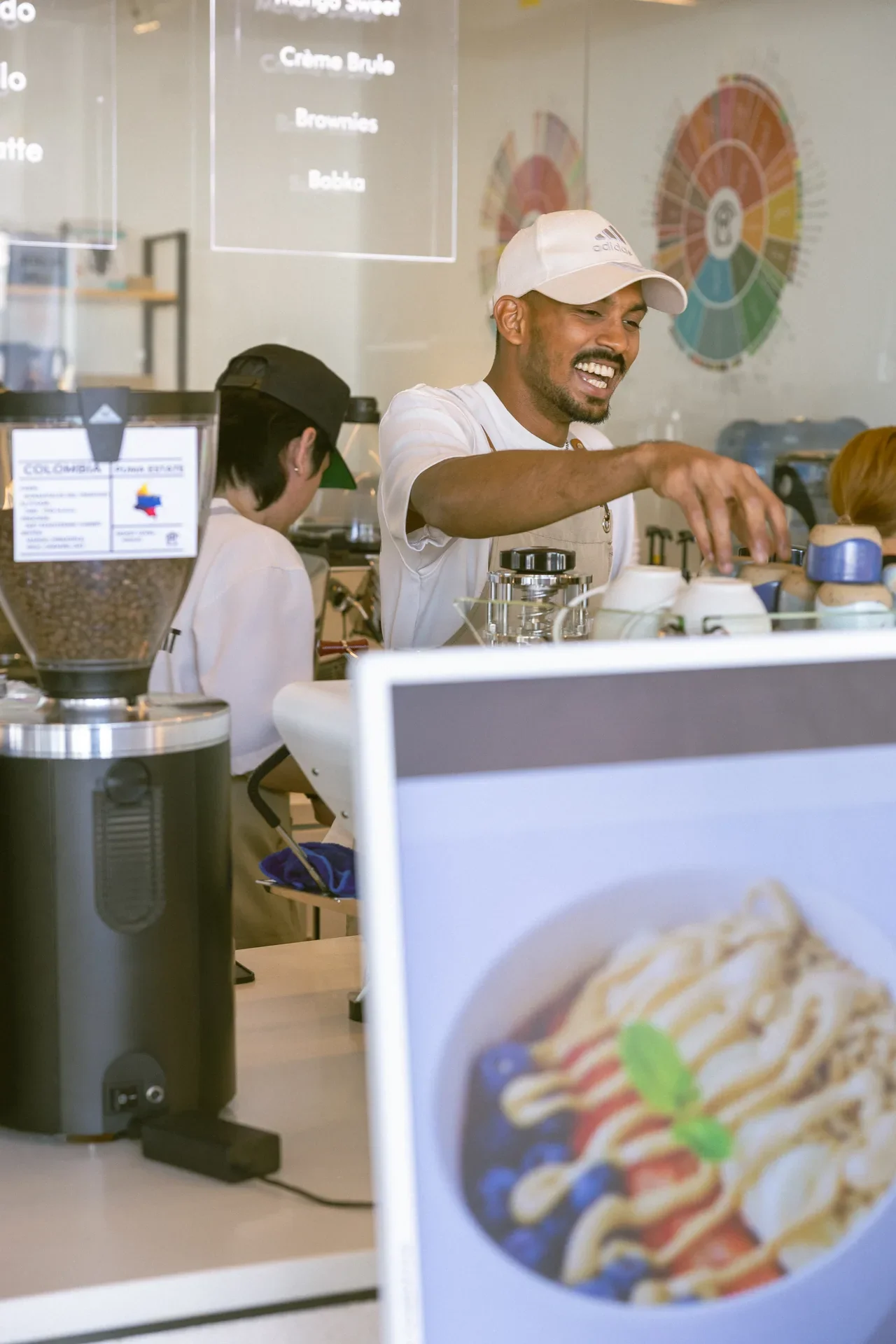 Smiling barista in a white cap and apron prepares coffee behind a cafe counter.