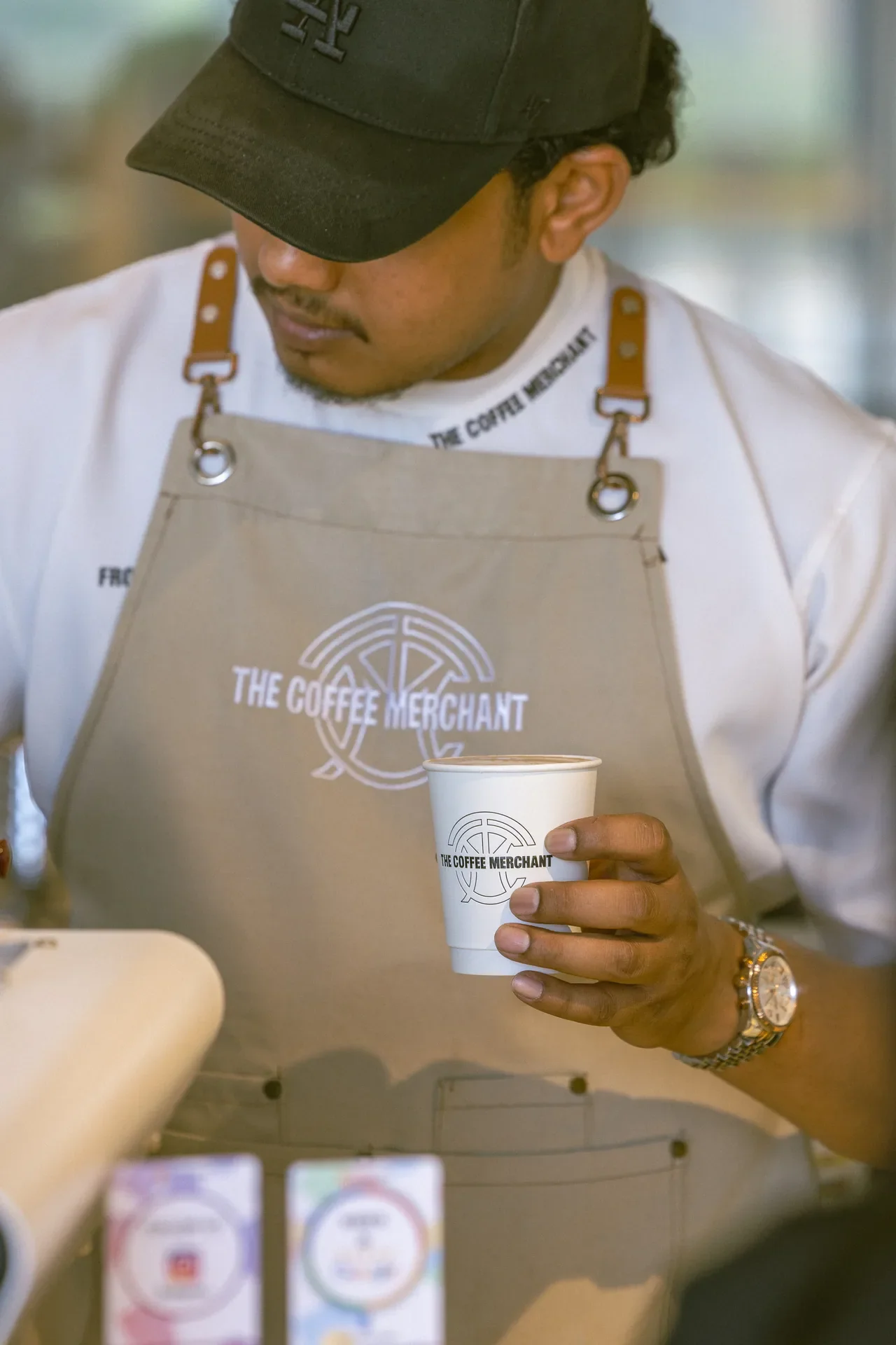 Barista wearing a beige apron with The Coffee Merchant logo, holding a paper coffee cup.