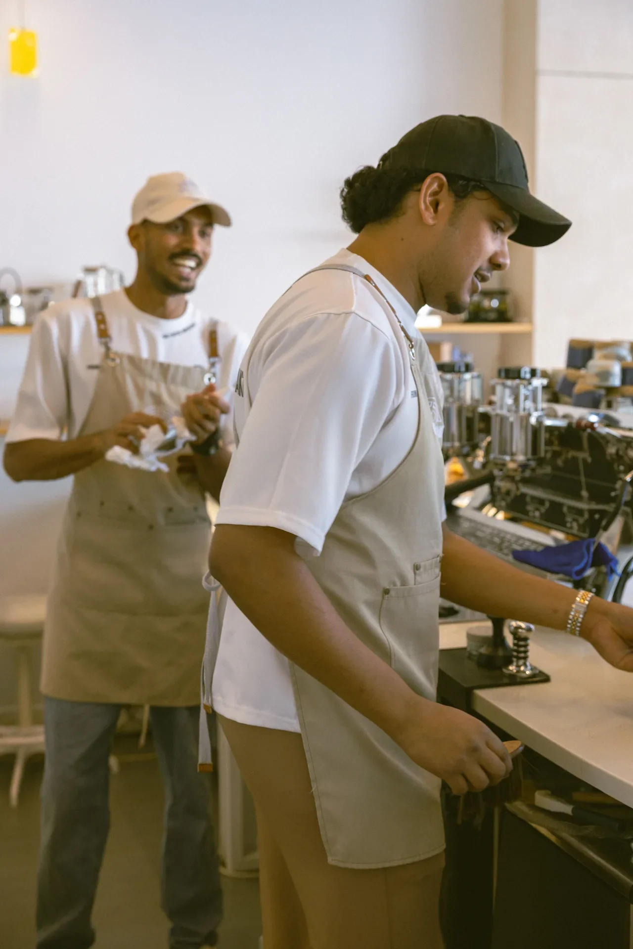 Two baristas in a cafe: one concentrating at an espresso machine, the other smiling in the background holding towels.