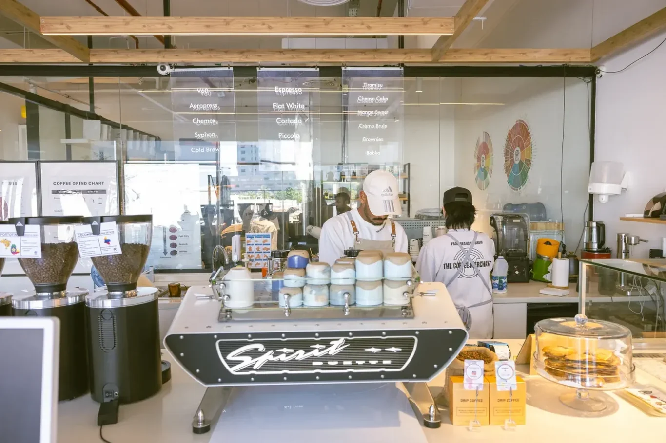 Barista behind a cafe counter preparing drinks, with an espresso machine and pastry display case.