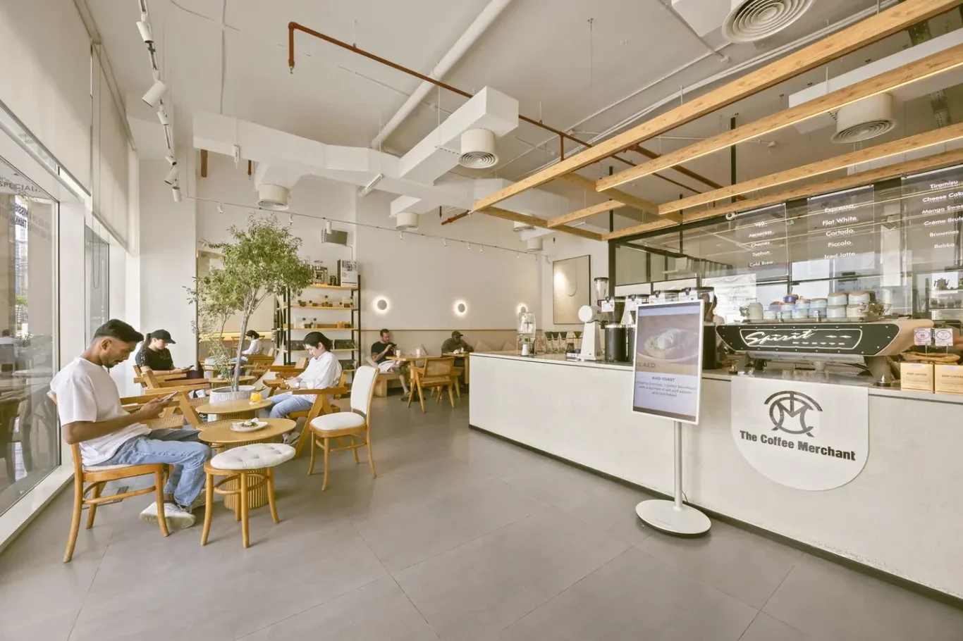 Bright cafe interior with wooden chairs, potted plant, and customers seated by large windows enjoying coffee and snacks.