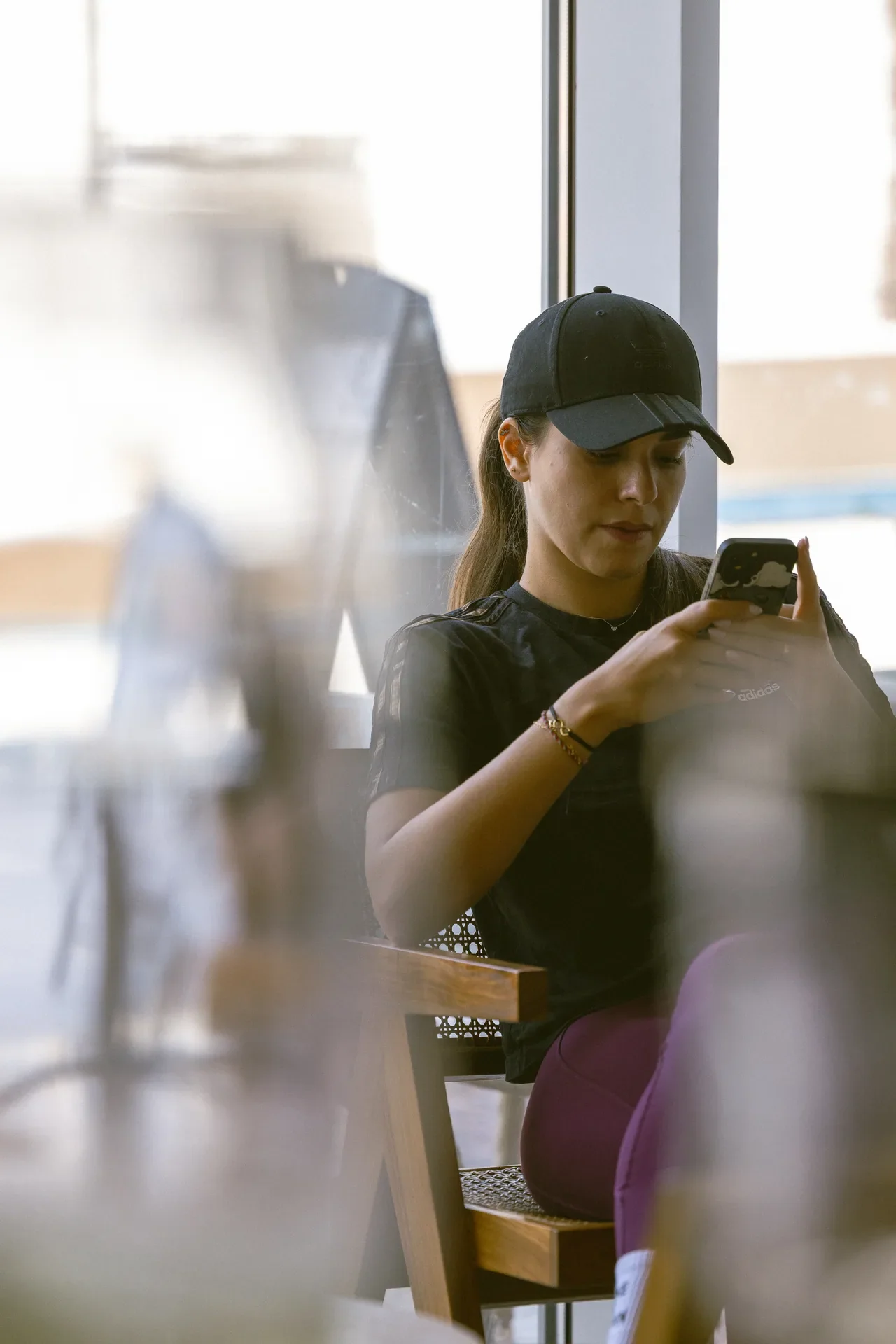 Young woman in a black cap and shirt sits at a cafe table, looking down at her smartphone by a window.