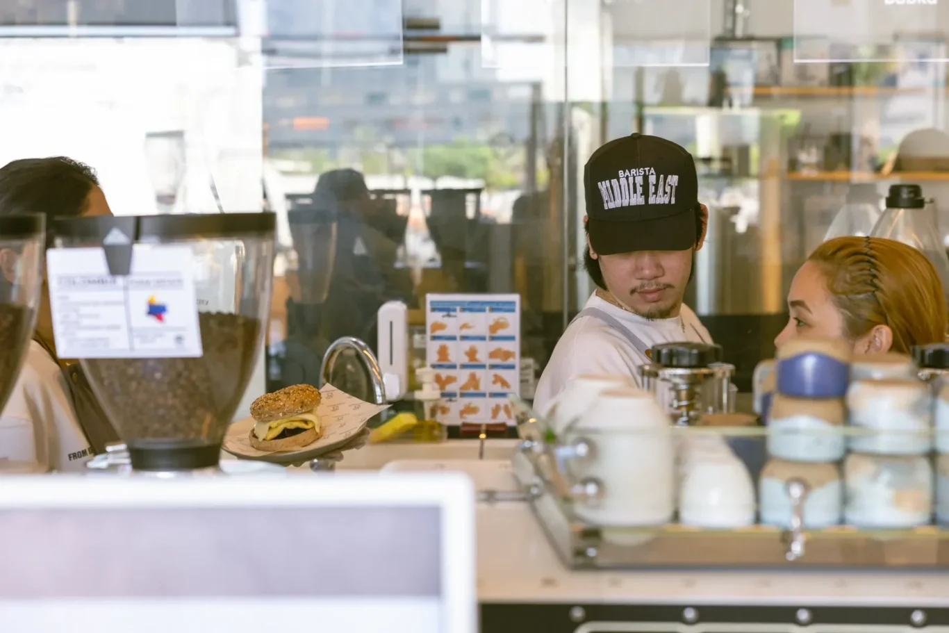 Barista wearing a cap that reads 'BARISTA MIDDLE EAST' behind a cafe counter, with a burger in the foreground.