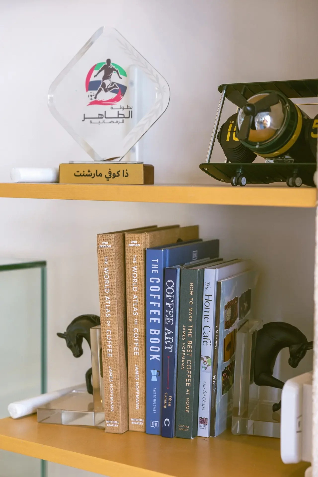 Glass diamond trophy with a soccer logo and Arabic inscription sits on a shelf beside a gold plaque with Arabic text and a row of coffee-table books.