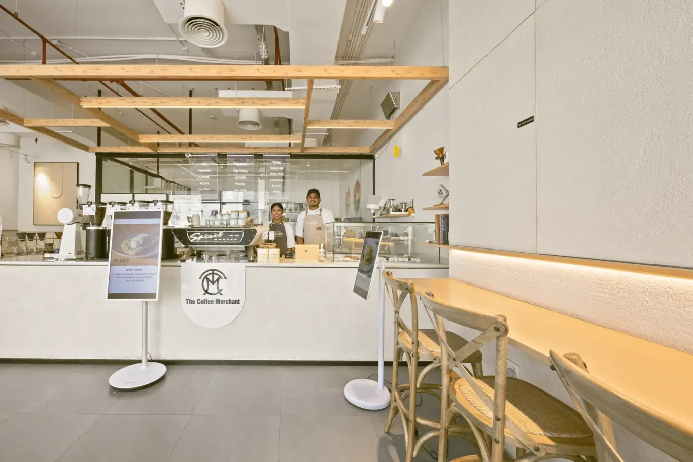 Coffee shop counter with two baristas behind glass, The Coffee Merchant logo on the front, wooden ceiling beams above