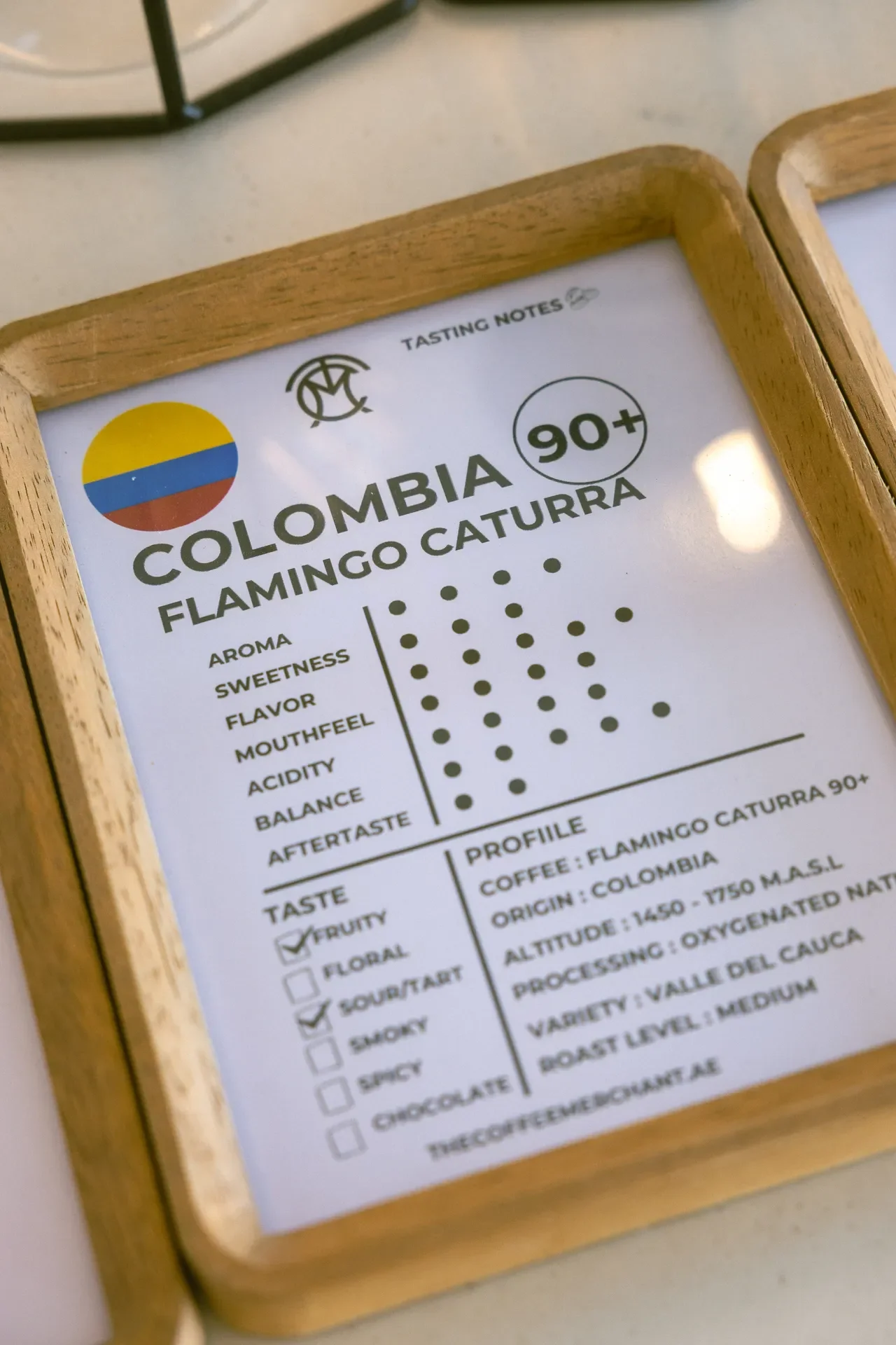 Tasting notes card for Colombia 90+ in a wooden frame, showing aroma, sweetness, flavor, mouthfeel, acidity, and balance checklists.
