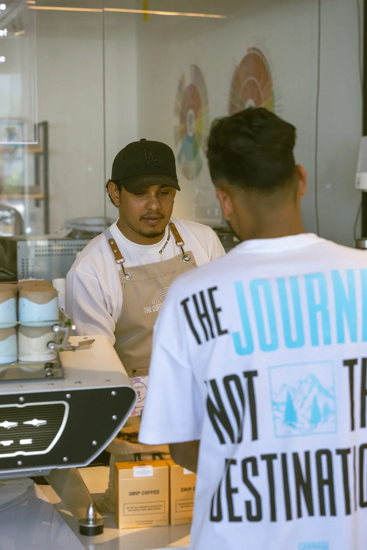 Barista in an LA cap and beige apron works at a coffee bar as a customer in a white T‑shirt with bold blue text stands nearby.