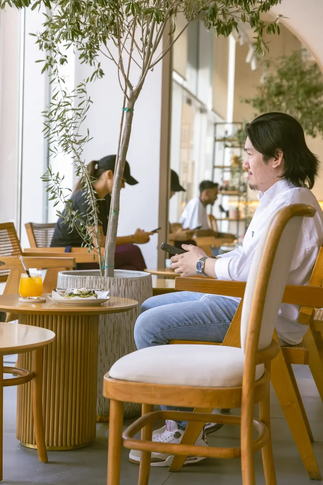 Man in a white shirt sits at a cafe table, looking at his phone, with a glass of orange juice and a plate of food nearby amid wooden furniture and plants in the background.