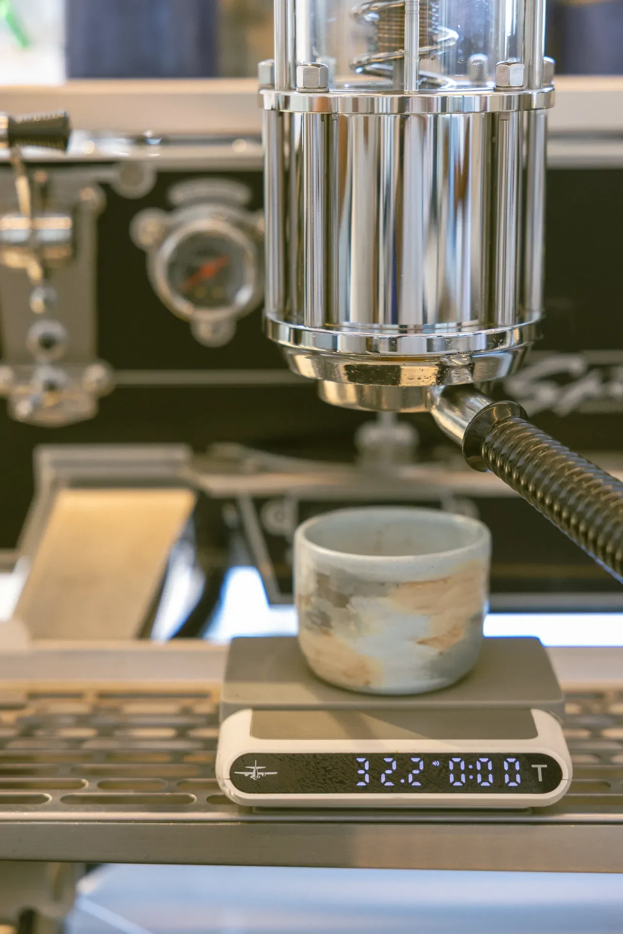 Close-up of a chrome espresso machine group head above a digital scale displaying 32.2 g and 0:00, with a mug nearby.