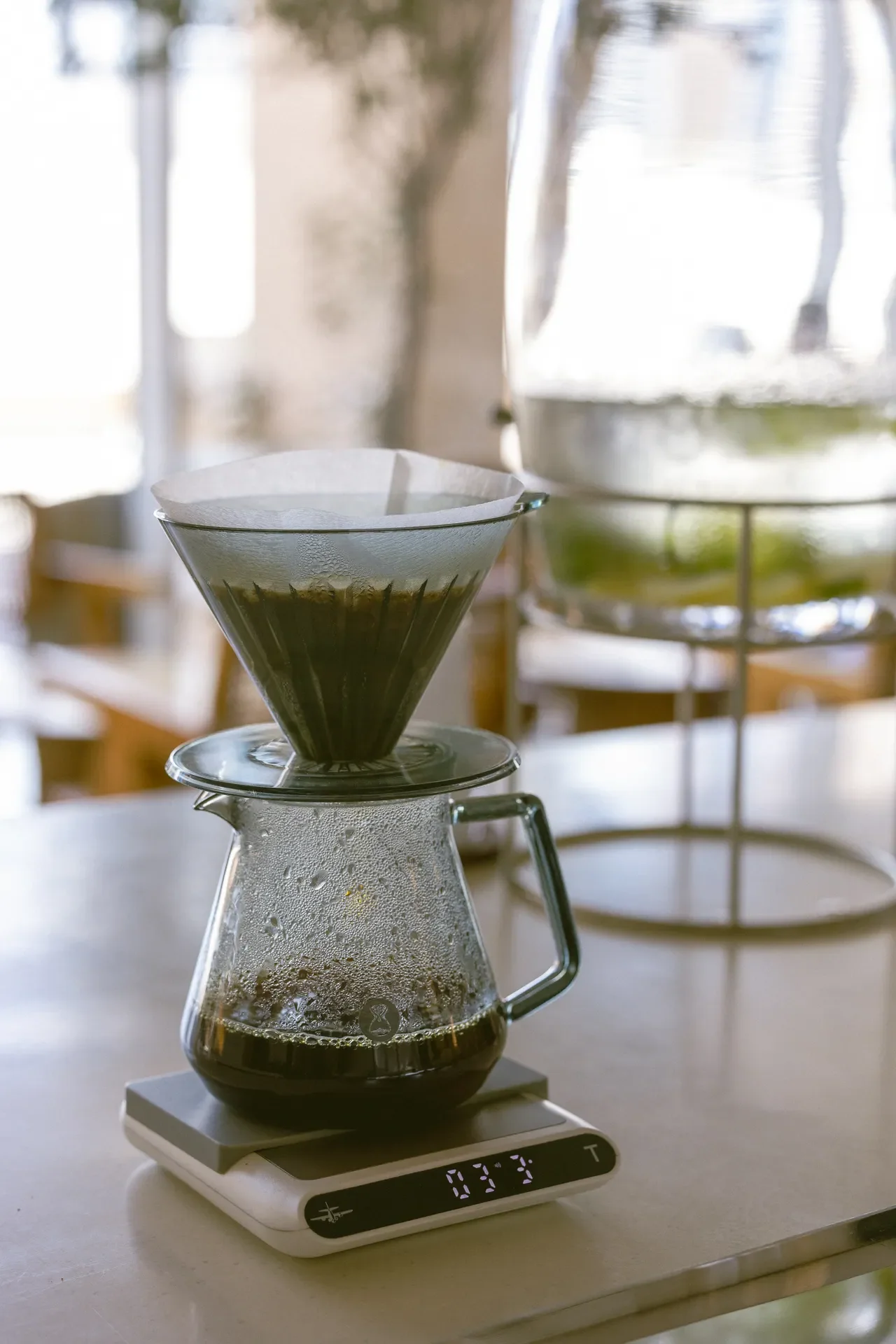 Pour-over coffee setup: a glass dripper with a paper filter perched on a glass carafe atop a digital scale on a kitchen counter.