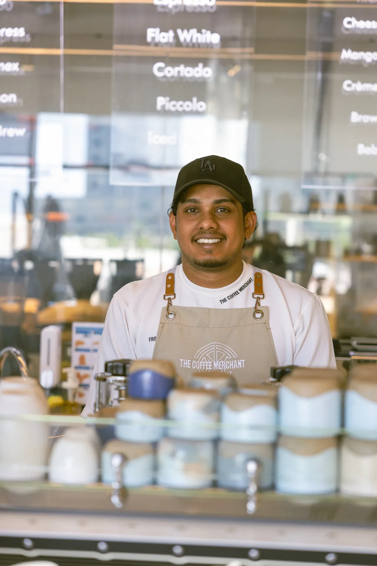 Smiling barista in a beige apron and black cap behind a coffee counter, with coffee jars in the foreground and a glass menu in the background.