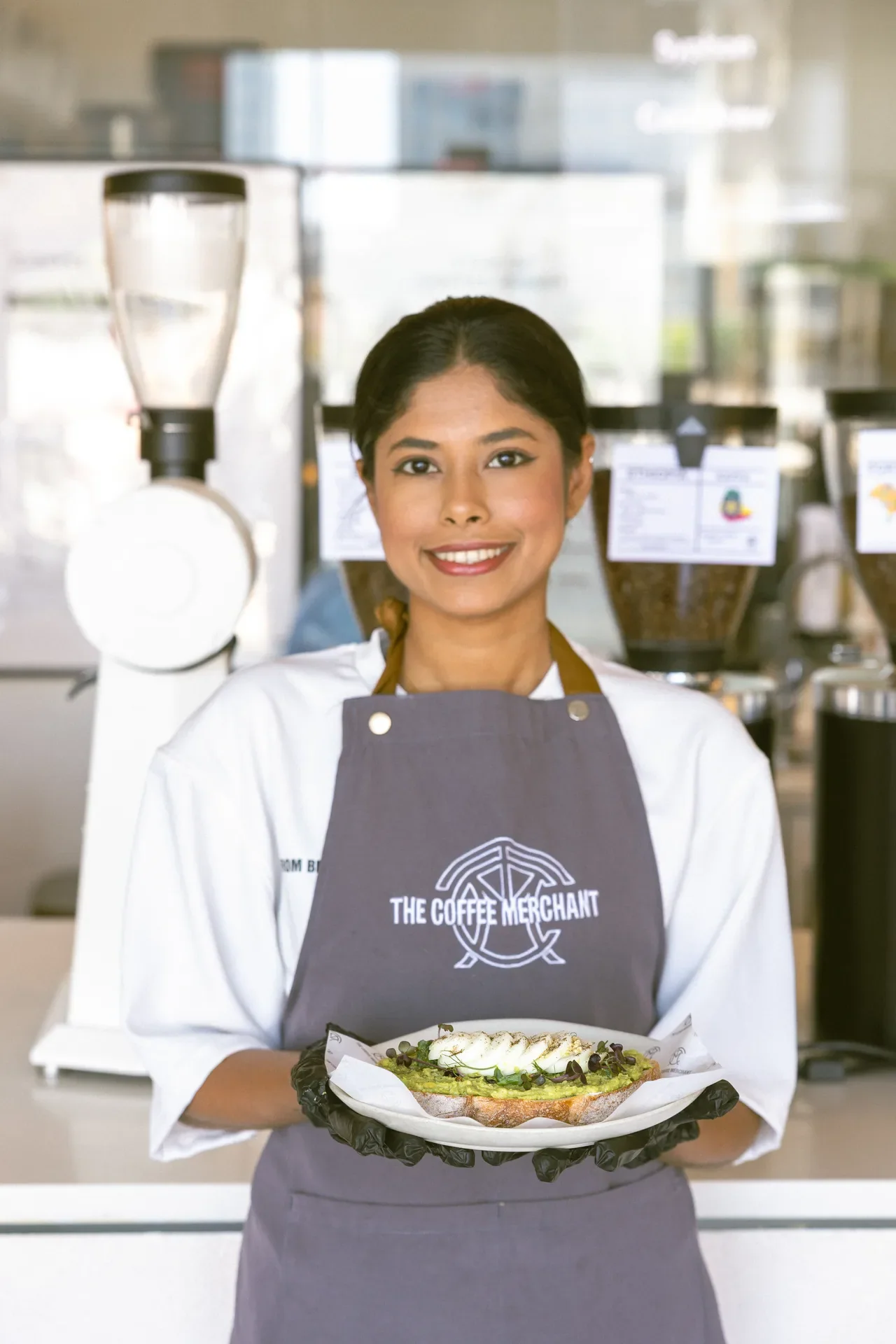Smiling barista in a gray apron presents an open-face avocado toast on a plate in a cafe setting