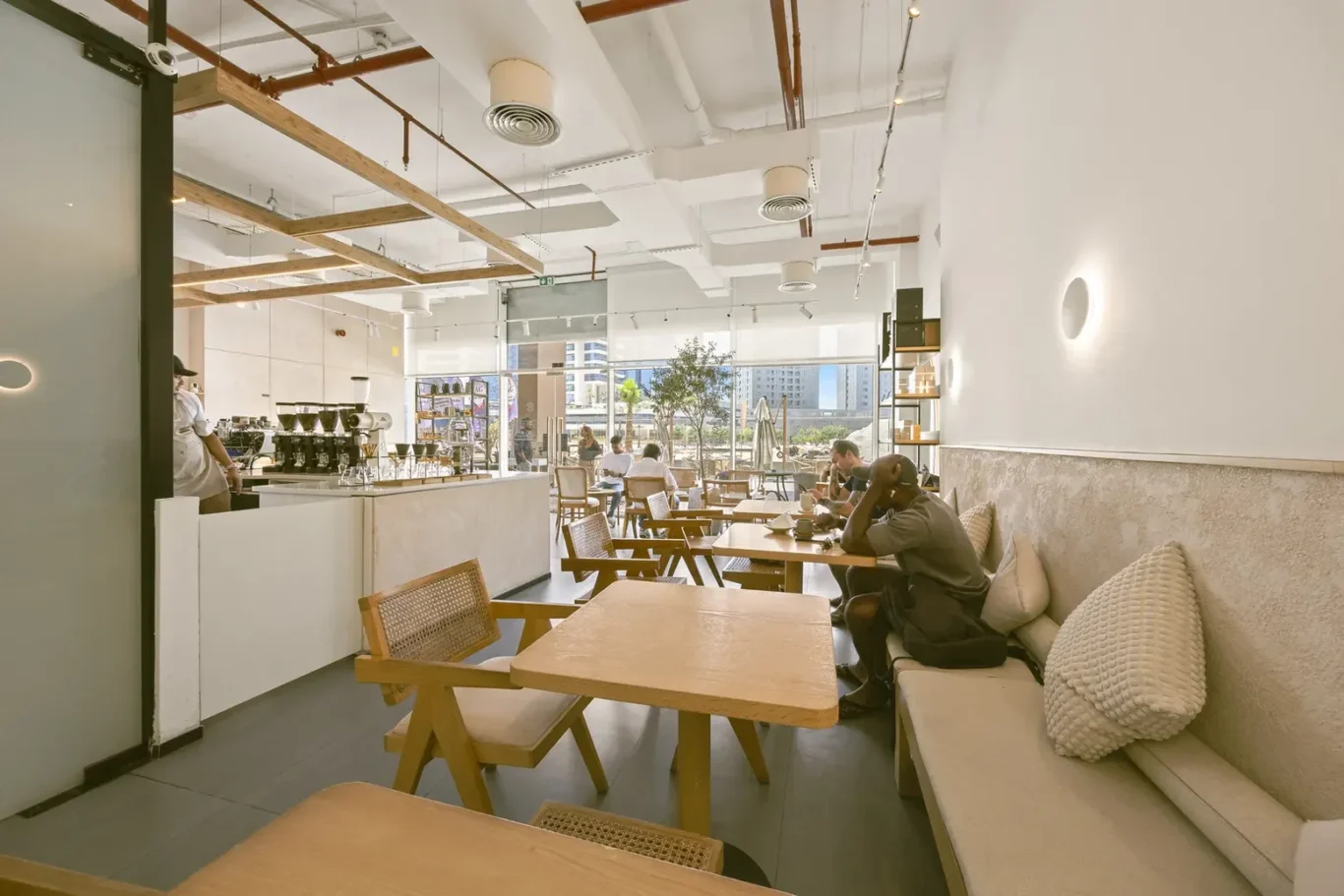 Bright modern cafe interior with wooden tables, bench seating, and customers near a large sunlit window.