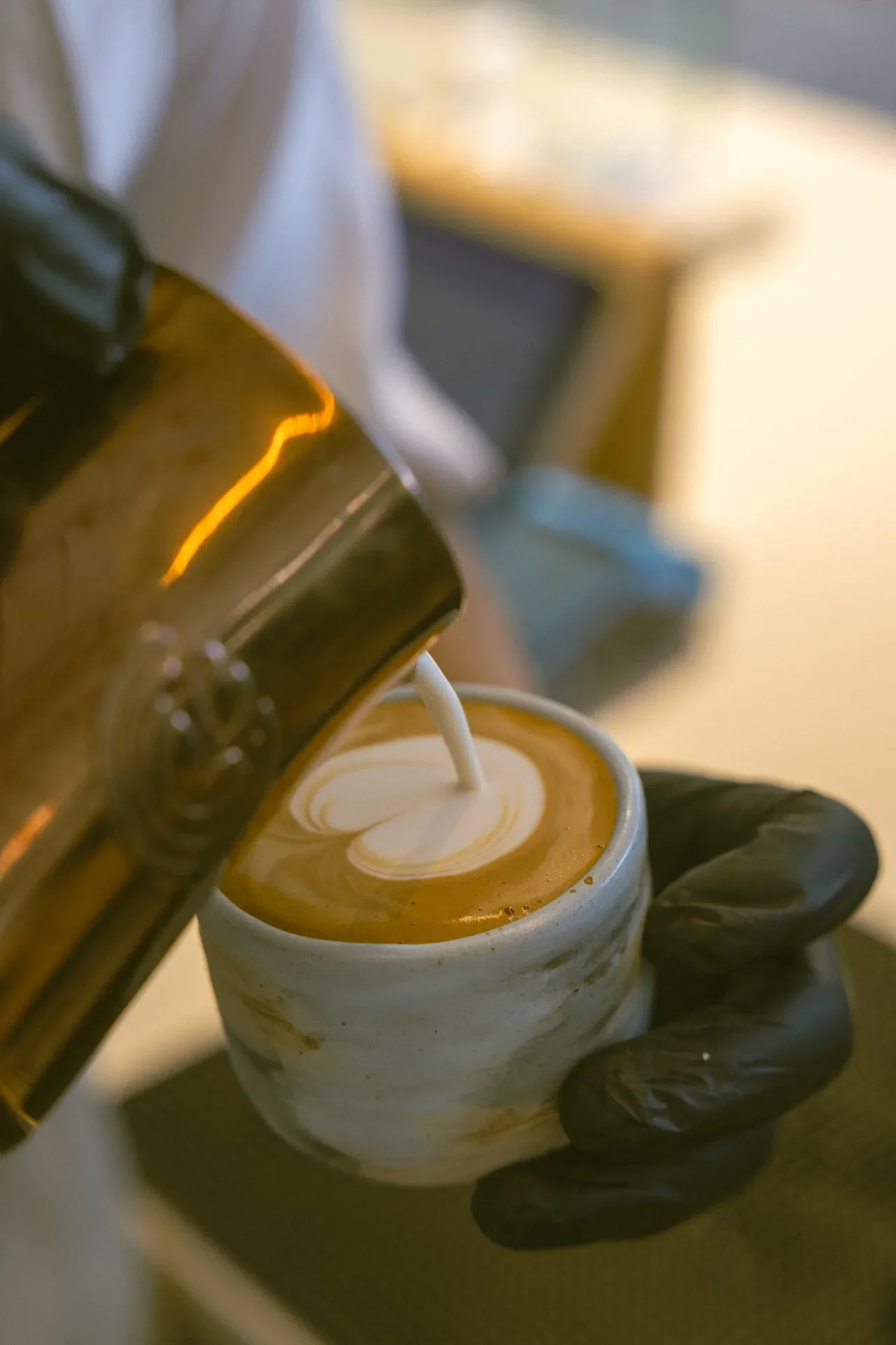 Barista wearing black gloves pours steamed milk into a cup of coffee, creating heart latte art on the surface.