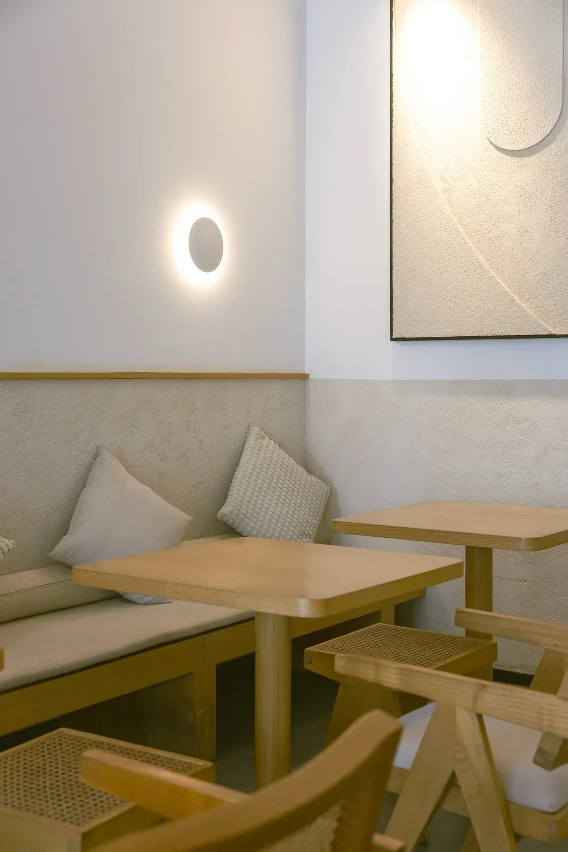 Cozy cafe seating with light wood tables, cushioned bench, and cream-colored walls with a round wall light and abstract artwork.