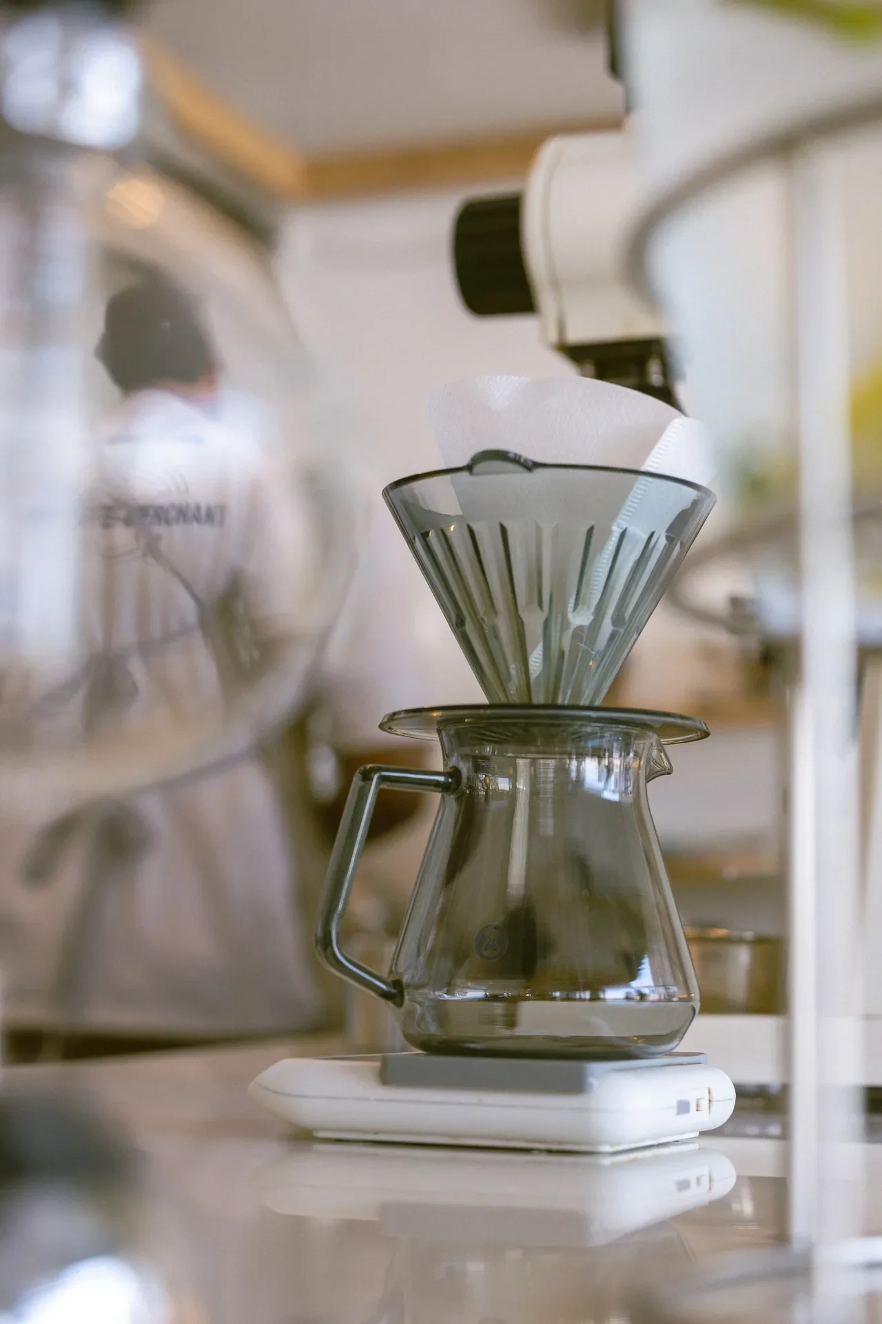 Pour-over coffee setup: cone filter in a dripper over a glass carafe on a scale, ready to brew in a bright kitchen.