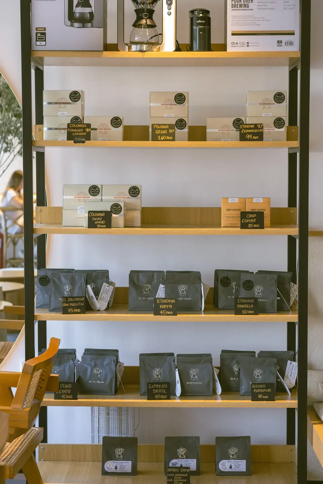 Display shelf of coffee products: rows of boxed and bagged coffee with chalkboard labels showing origins and prices (e.g., Colombia, Ethiopia, DRIP coffee) in a shop setting.