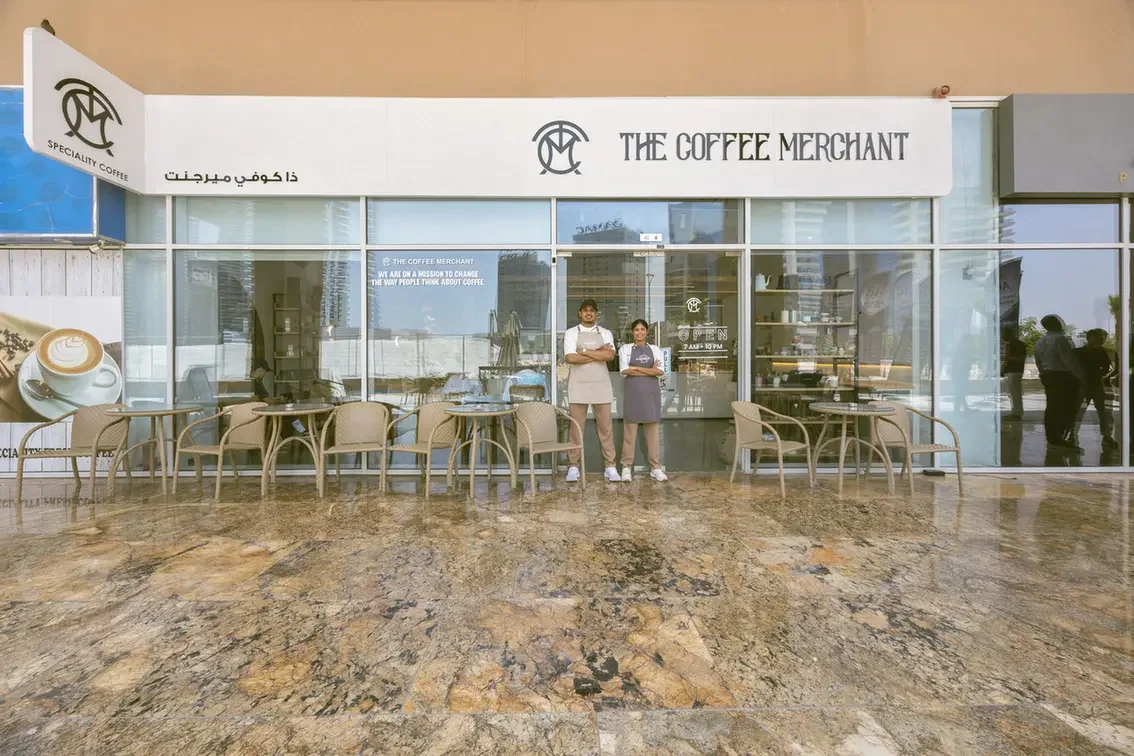 The Coffee Merchant cafe storefront with two staff members posing outside, glass windows and outdoor tables.