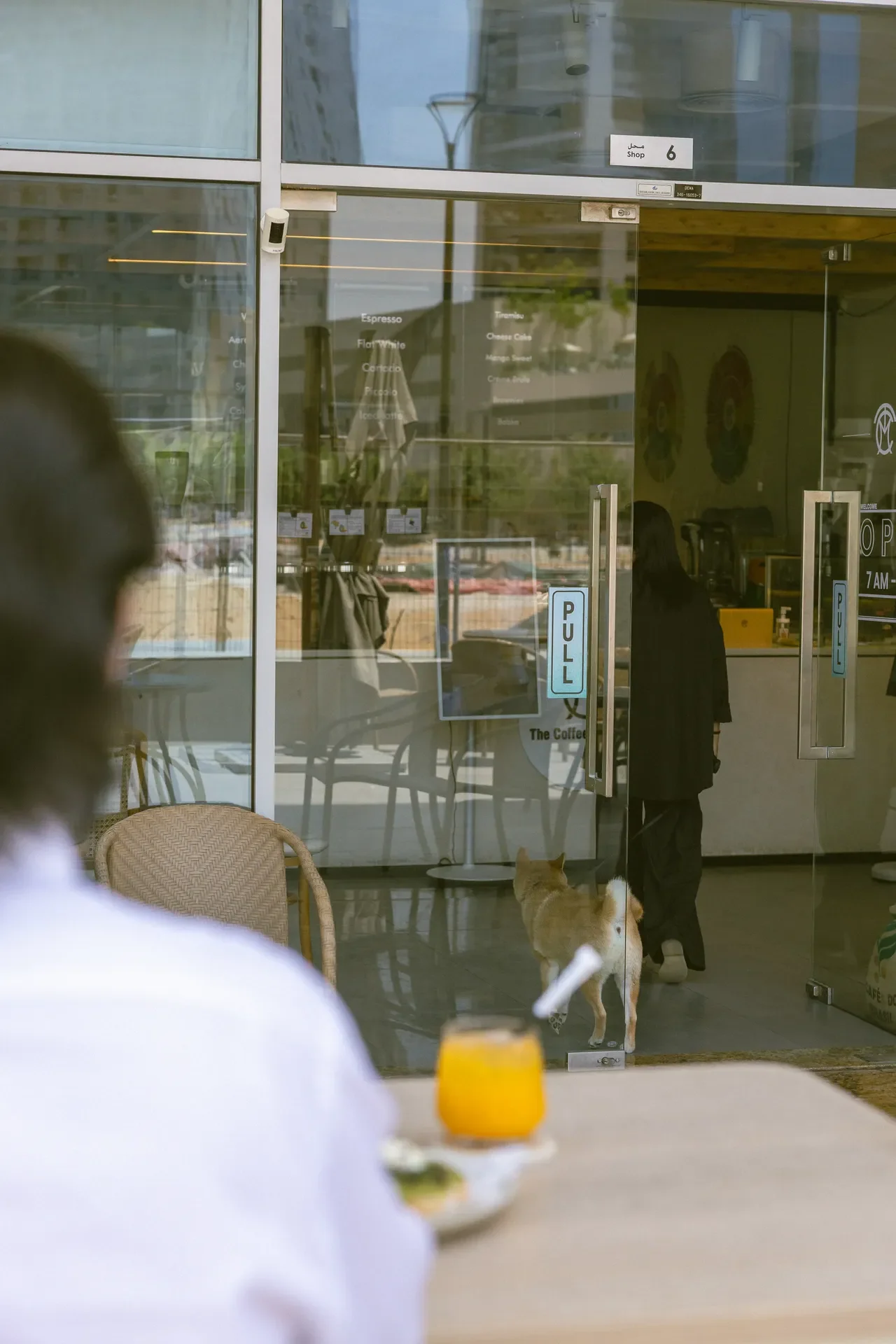 Cafe interior with glass doors; there is a light-colored dog standing near a person in black, foreground shows a yellow drink on a table.