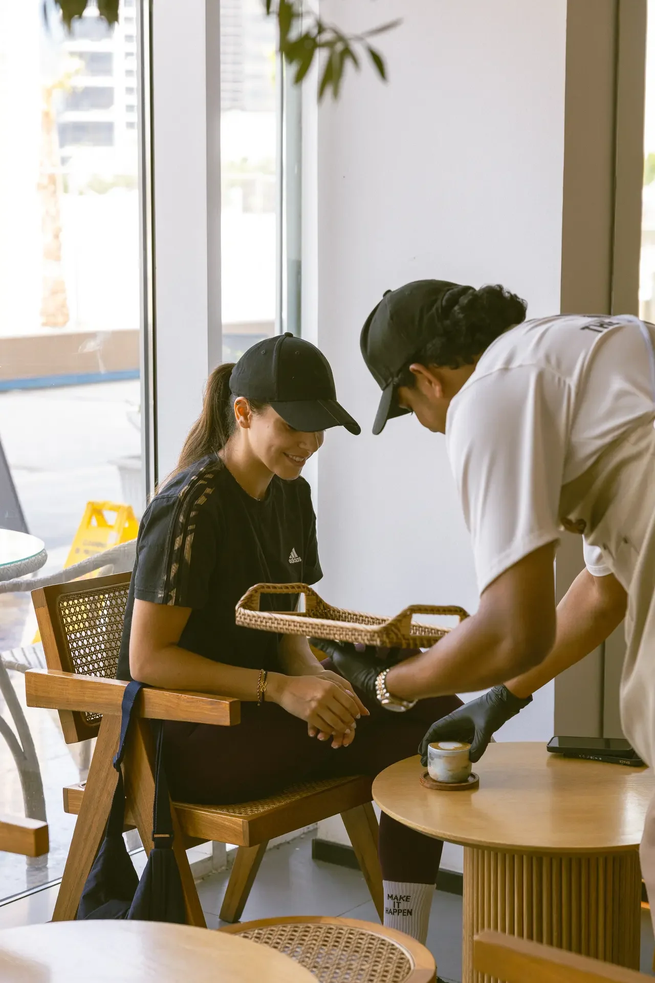 Woman wearing a black cap sits in a wooden chair while a worker with a cap leans over her hands for a manicure in a bright, cafe-like setting.