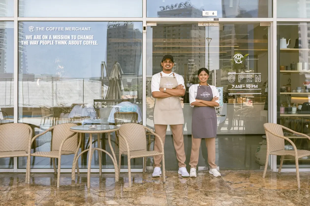 Two cafe staff stand with arms crossed outside a glass-front coffee shop, ready to serve customers.