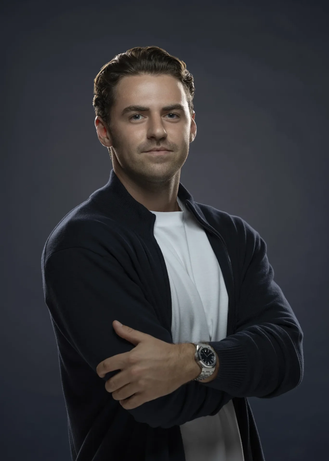 Professional headshot of a man with arms crossed, wearing a white shirt and navy cardigan, against a dark studio backdrop.